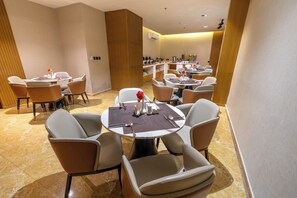 A modern dining area with round tables and cushioned chairs, a wooden accent wall, and a bar area in the background. - Al MuhaidbResidence Al Takhasossi 1 - Economic (Riyadh)
