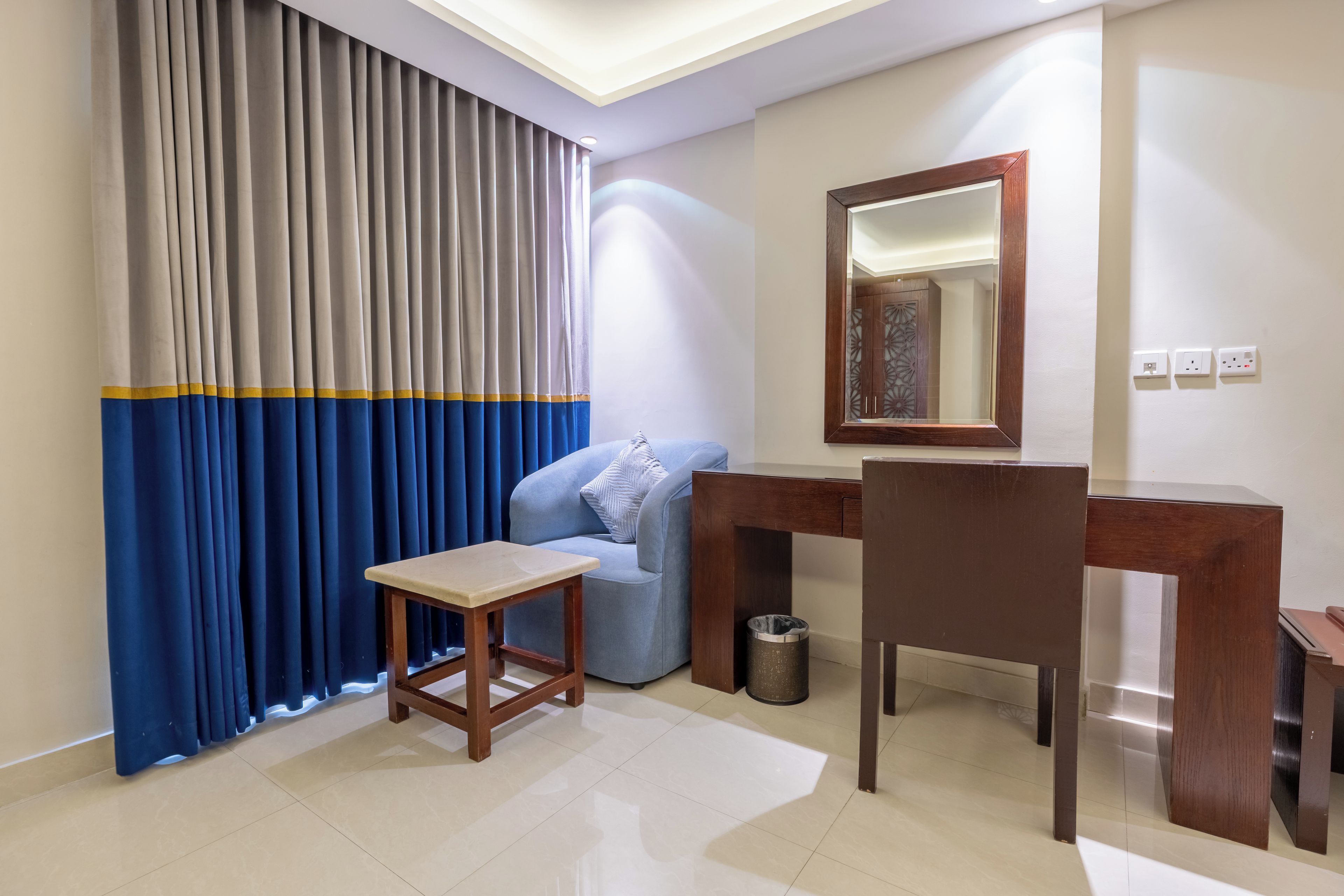 A hotel room with a bed, a desk, a chair, a small table, a mirror, a waste bin, and a window with curtains.
