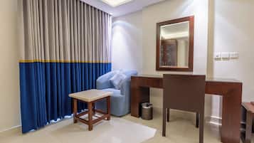 A hotel room with a bed, a desk, a chair, a small table, a mirror, a waste bin, and a window with curtains.