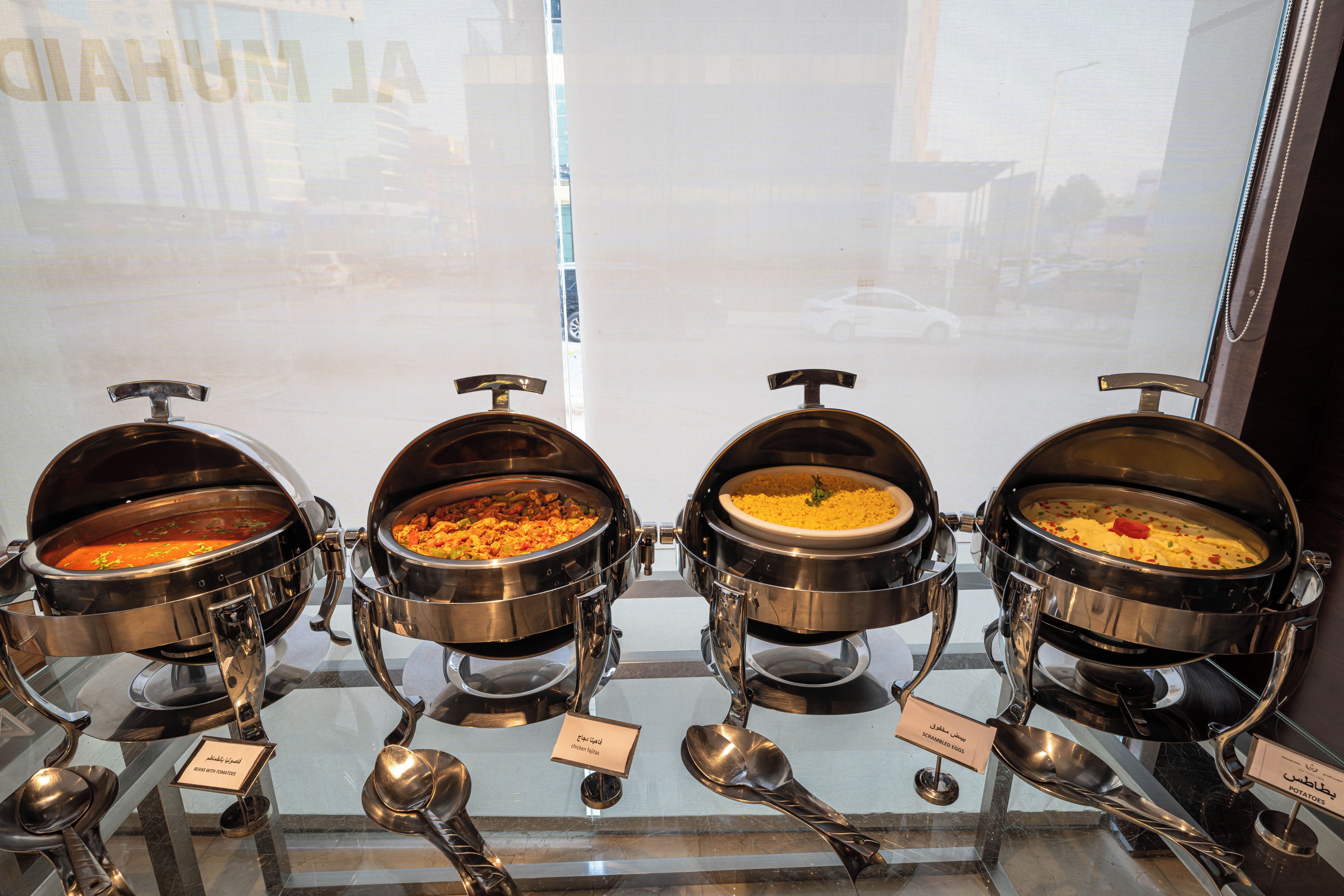 A buffet with four different dishes, each in a separate serving station with a spoon and a label indicating the dish name.