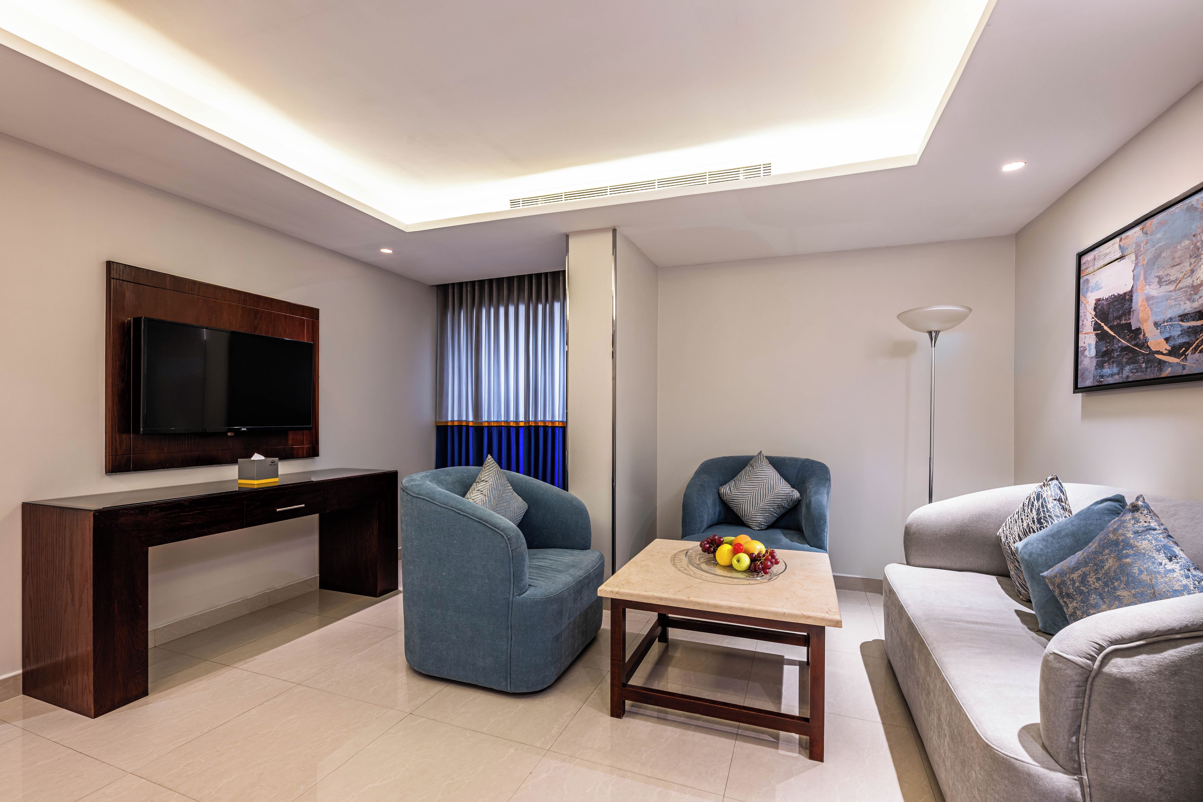A modern living room with a flat-screen TV, a dark wooden desk, a blue armchair, a round coffee table with fruit, and a grey sofa.