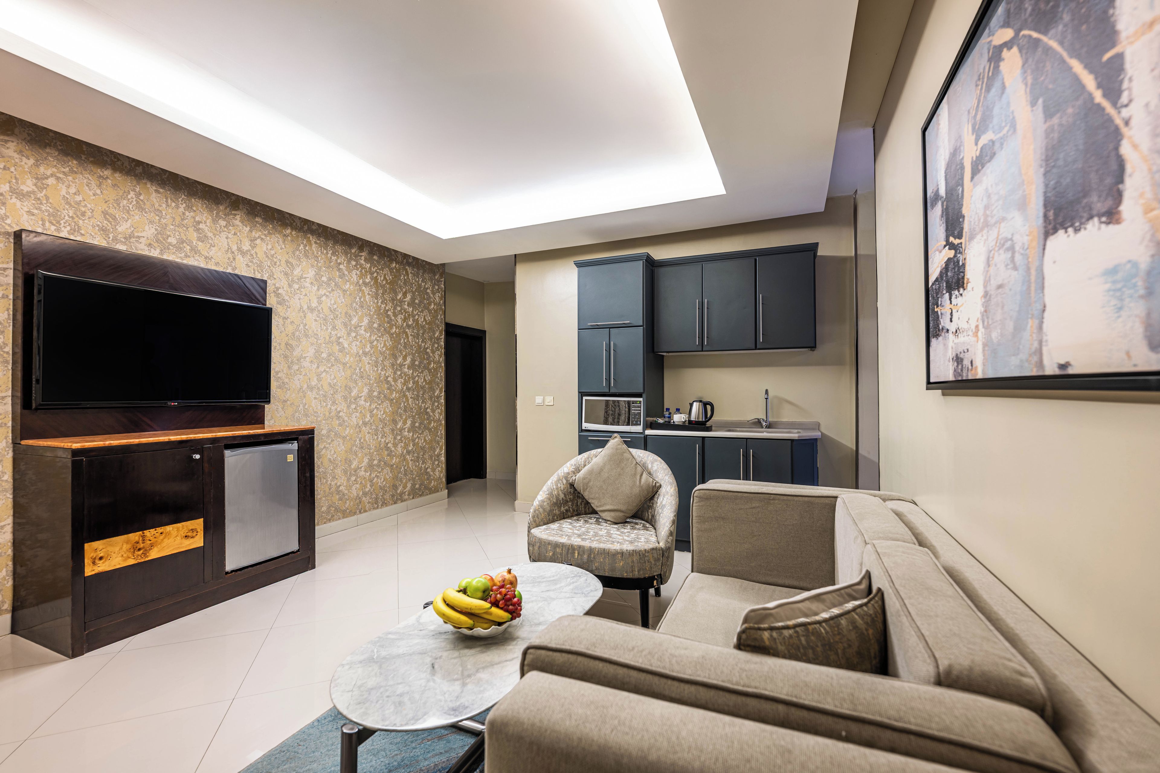 A modern living room with a flat-screen TV, a sofa, a coffee table with fruit, and a kitchen area in the background.