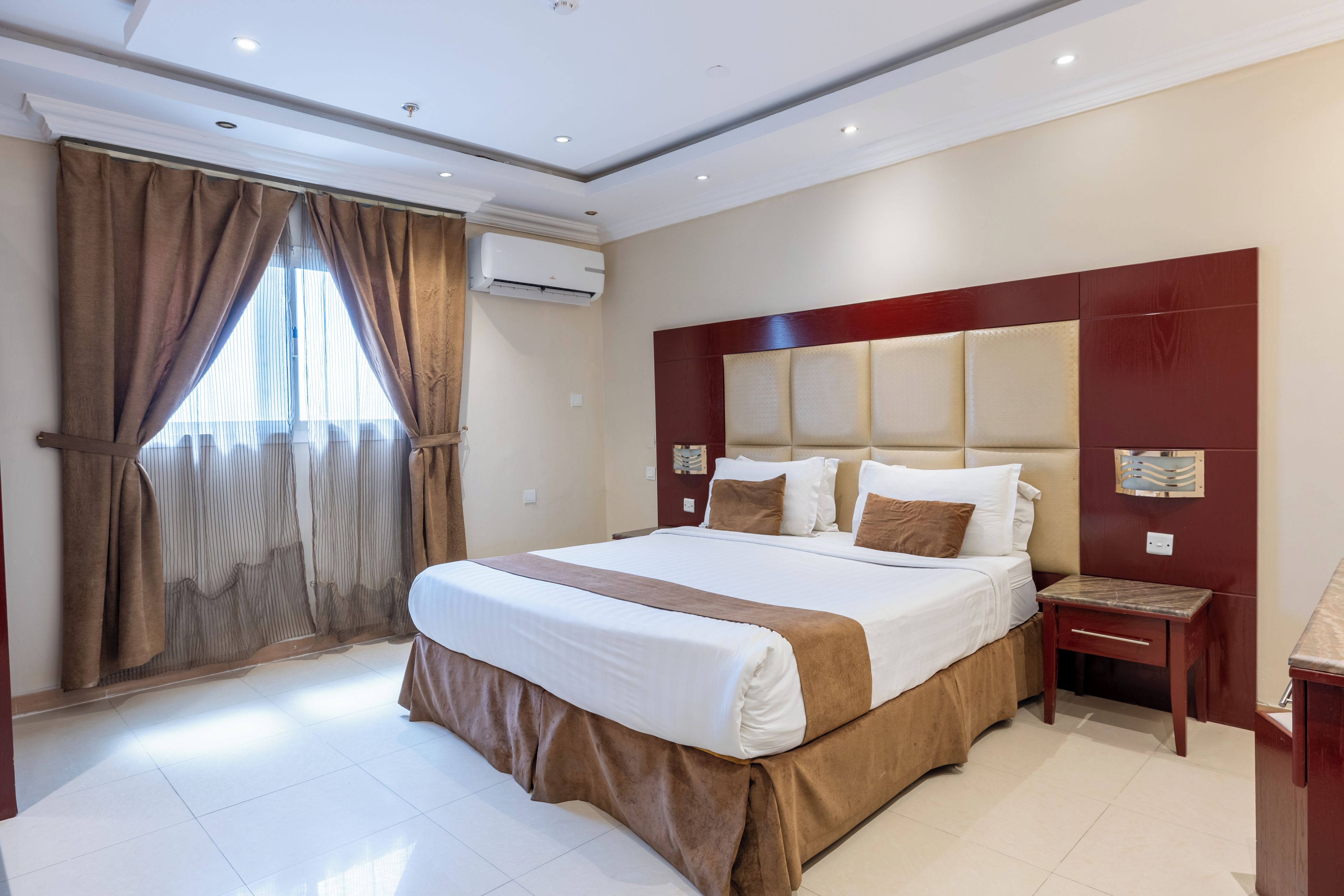 A hotel room with a large bed, a nightstand, a wall-mounted air conditioner, and a window with curtains.
