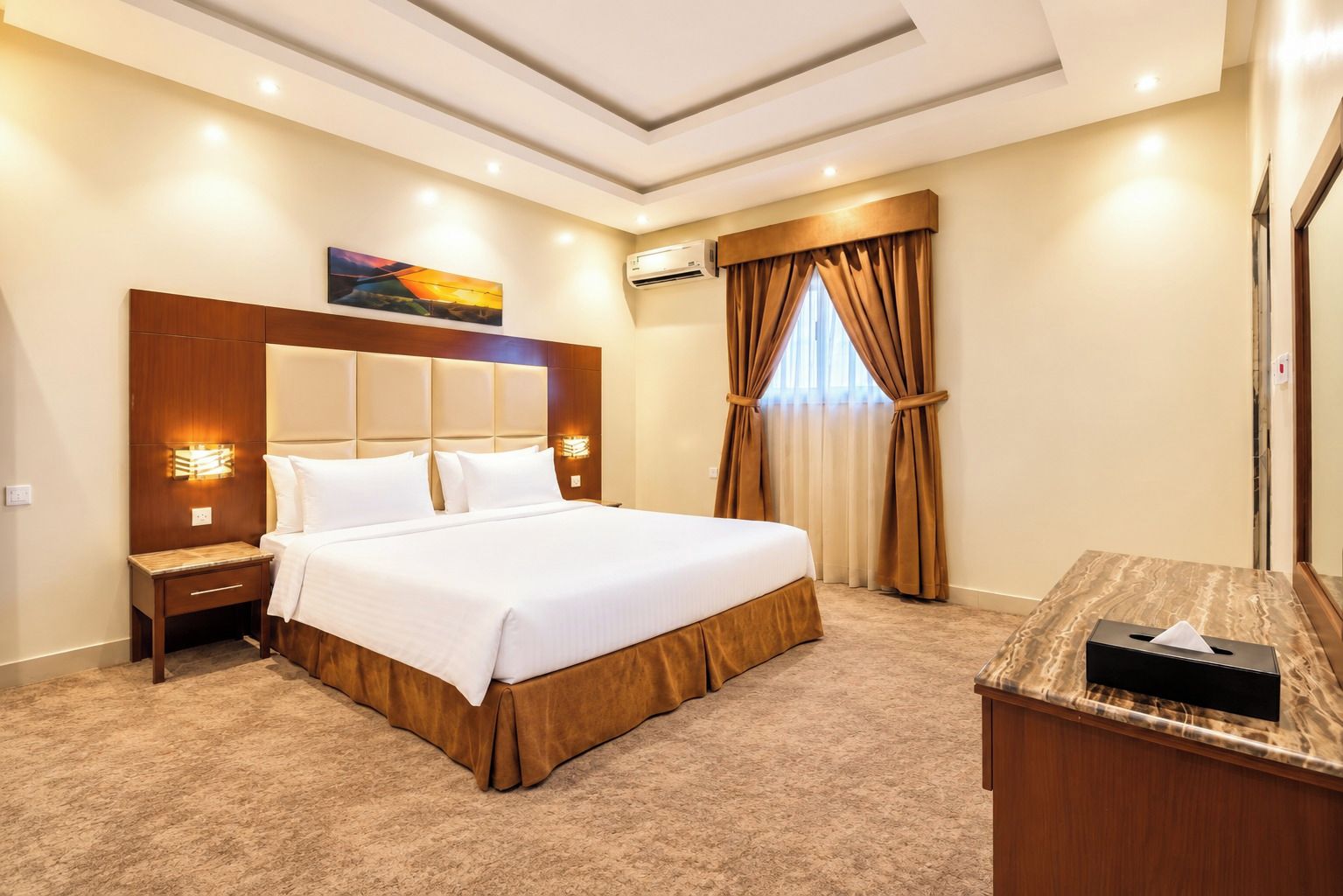A hotel room with a large bed, a bedside table, an air conditioning unit, a window with curtains, and a painting on the wall.