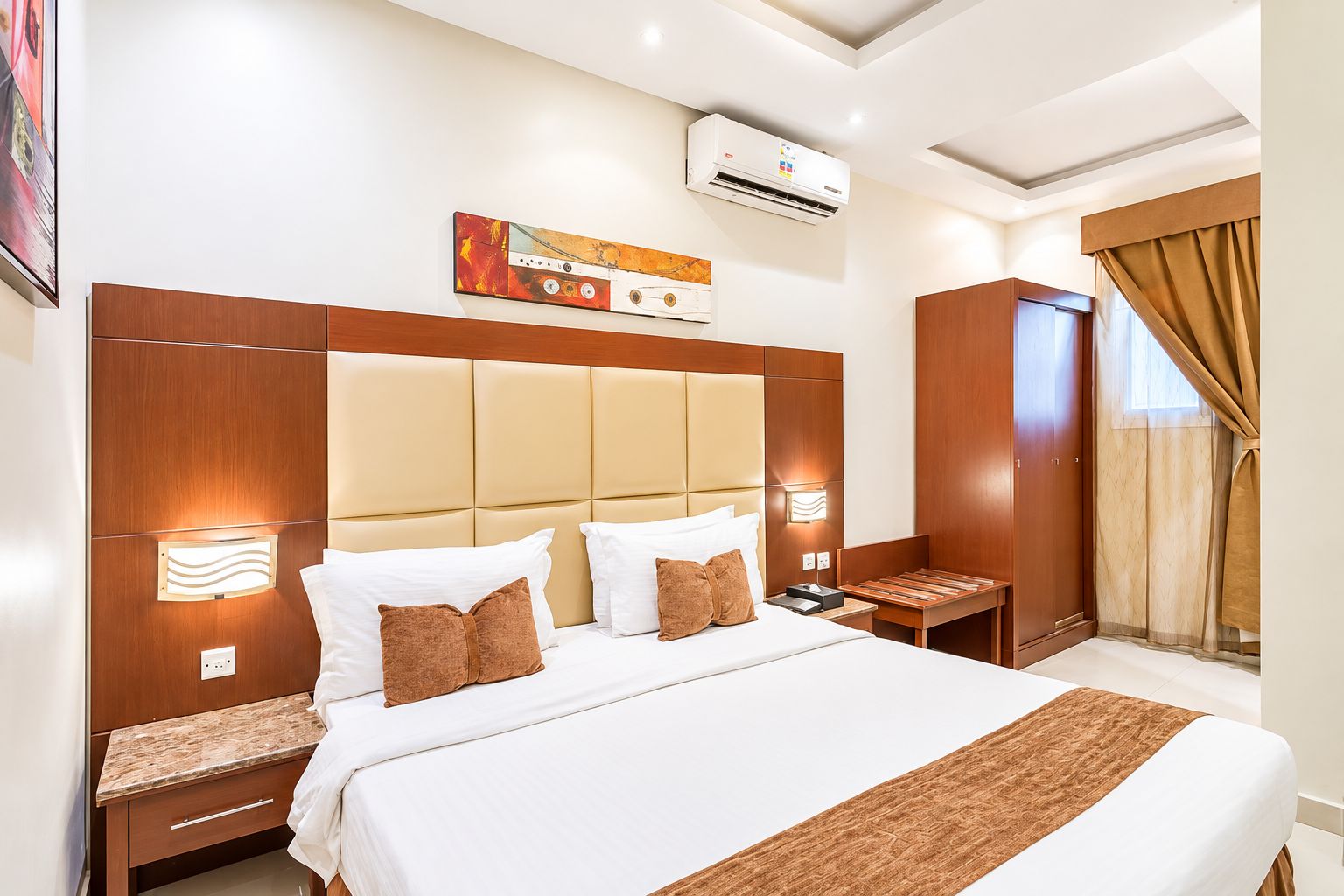 Ein Hotelzimmer mit einem großen Bett, einem hölzernen Kopfteil, einem Nachttisch, einer Fensterbank, einer Klimaanlage und einem Fenster mit Vorhängen.