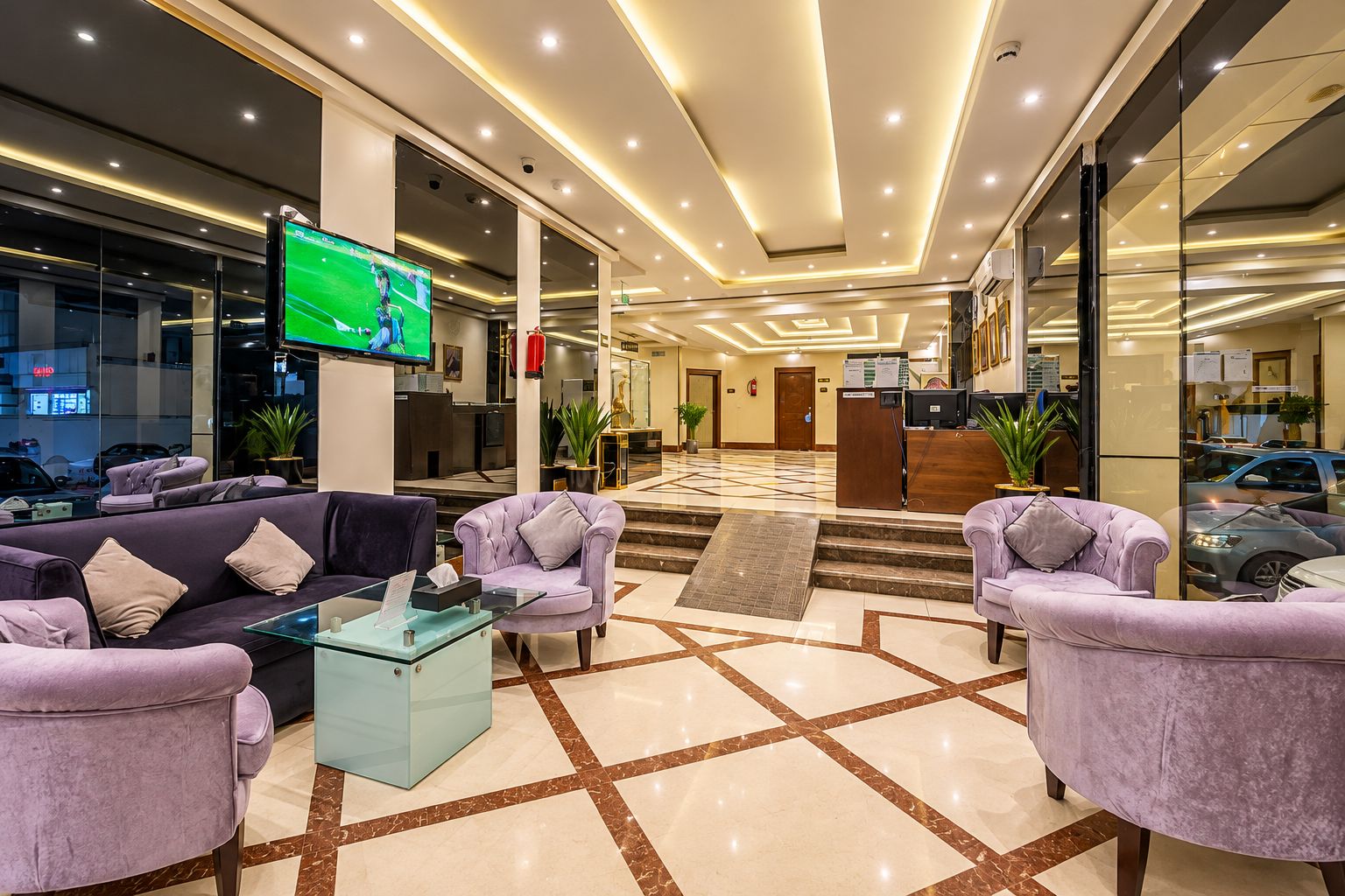 A hotel lobby with a reception desk, seating area with sofas and armchairs, a television screen, and a patterned floor.