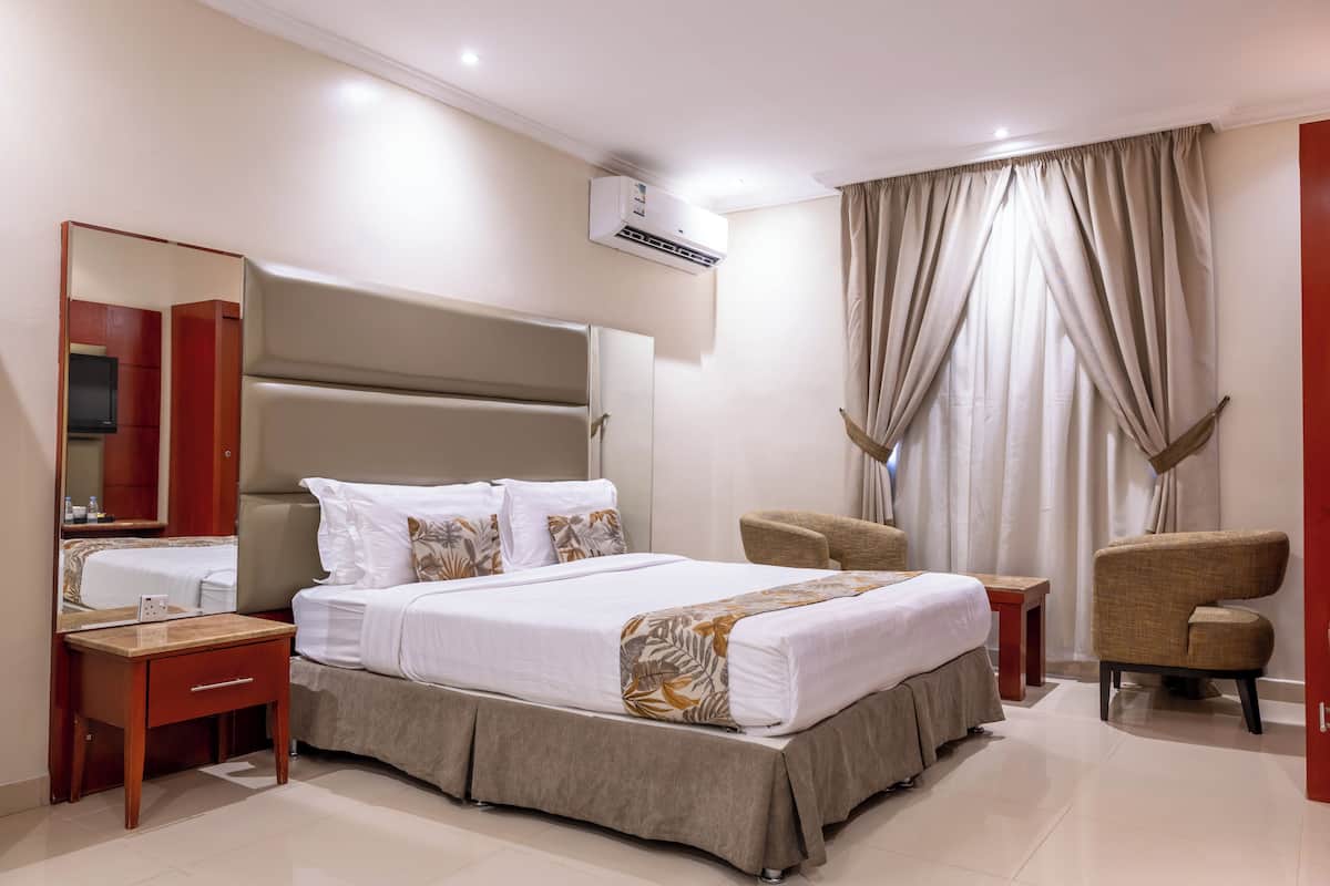 A hotel room with a large bed, a bedside table, a chair, a mirror, and an air conditioning unit.