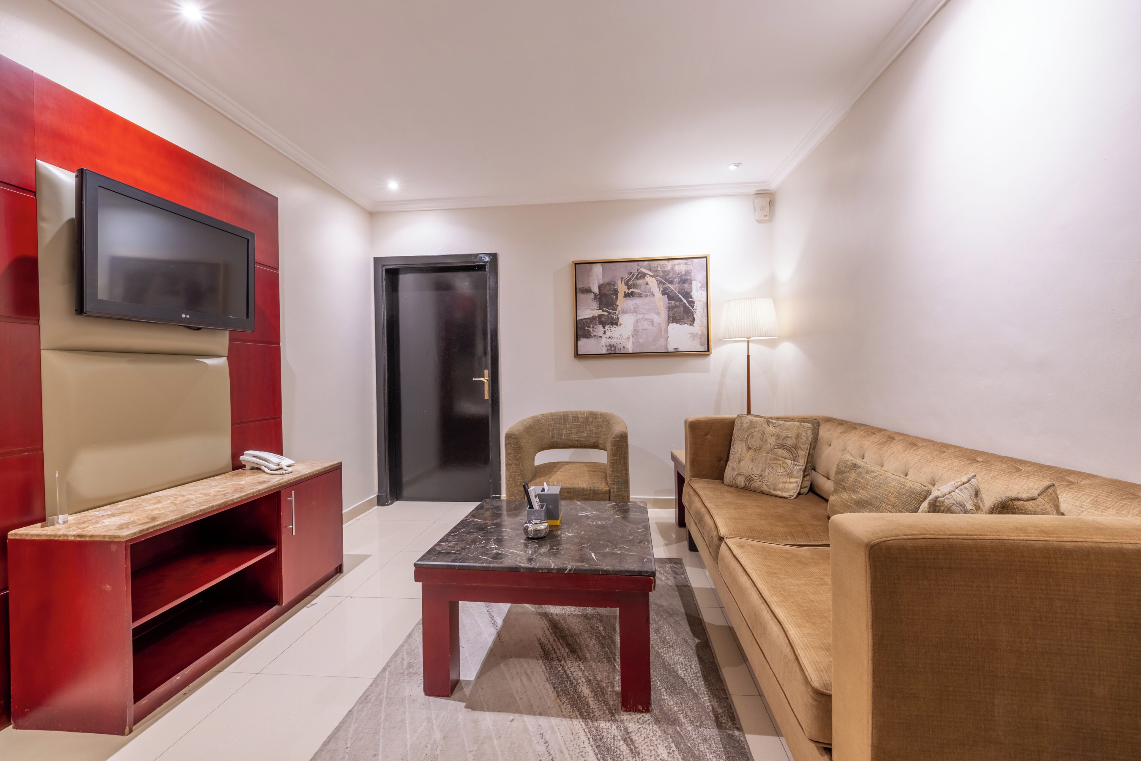A living room with a sofa, a coffee table, a TV mounted on a red wall, a door, and a framed picture on the wall.