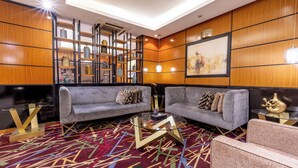 Una habitación de hotel moderna con dos sofás grises, una mesa de centro de cristal, alfombra estampada, paneles de madera y una obra de arte enmarcada.