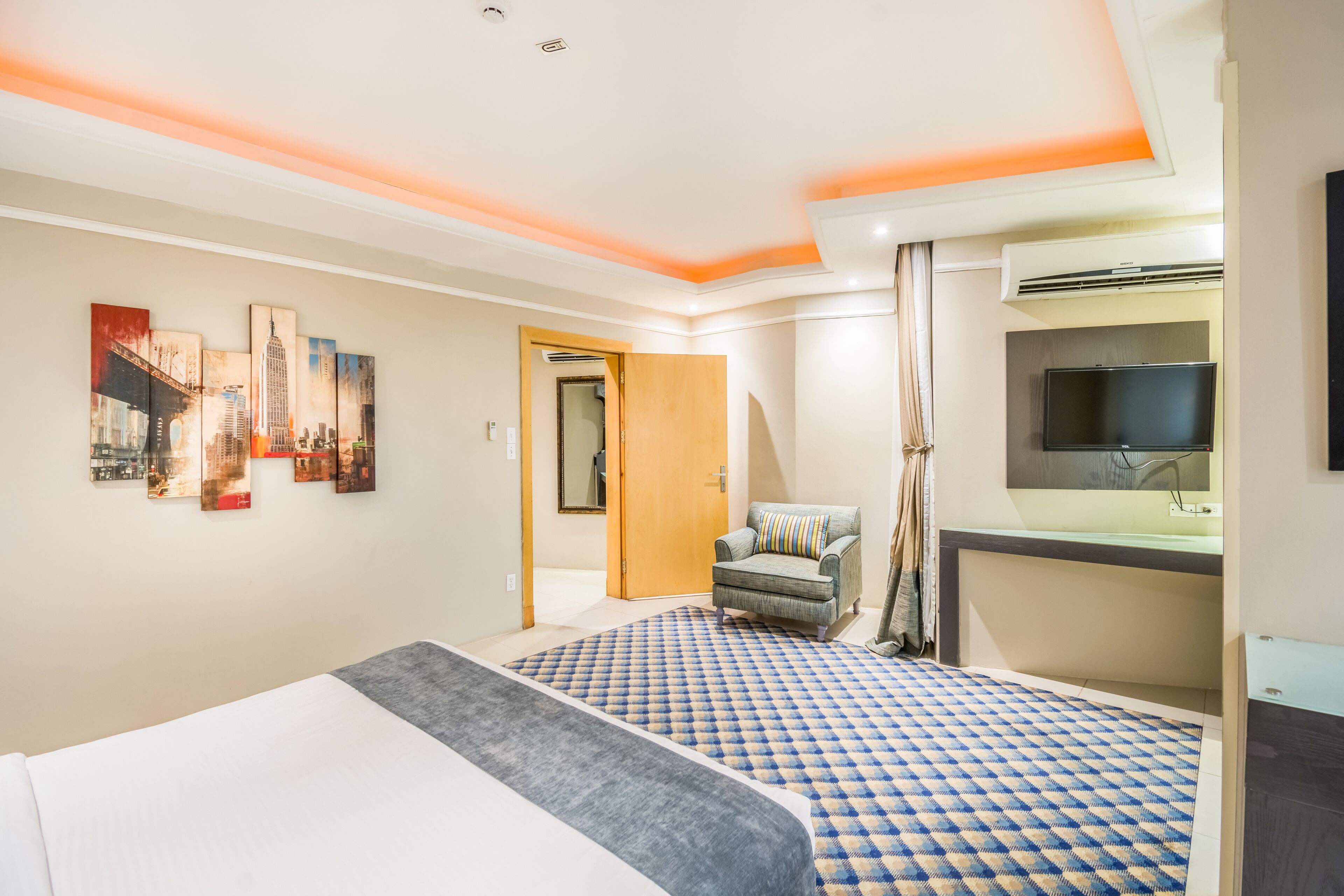 A hotel room with a bed, a desk with a flat-screen TV, a chair, and a bathroom with a mirror and sink.