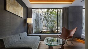 Ein modernes Wohnzimmer mit einem grauen Sofa, einem Glastisch und einem braunen Sessel. Ein großes Fenster bietet Blick auf Bäume und einen Balkon.
