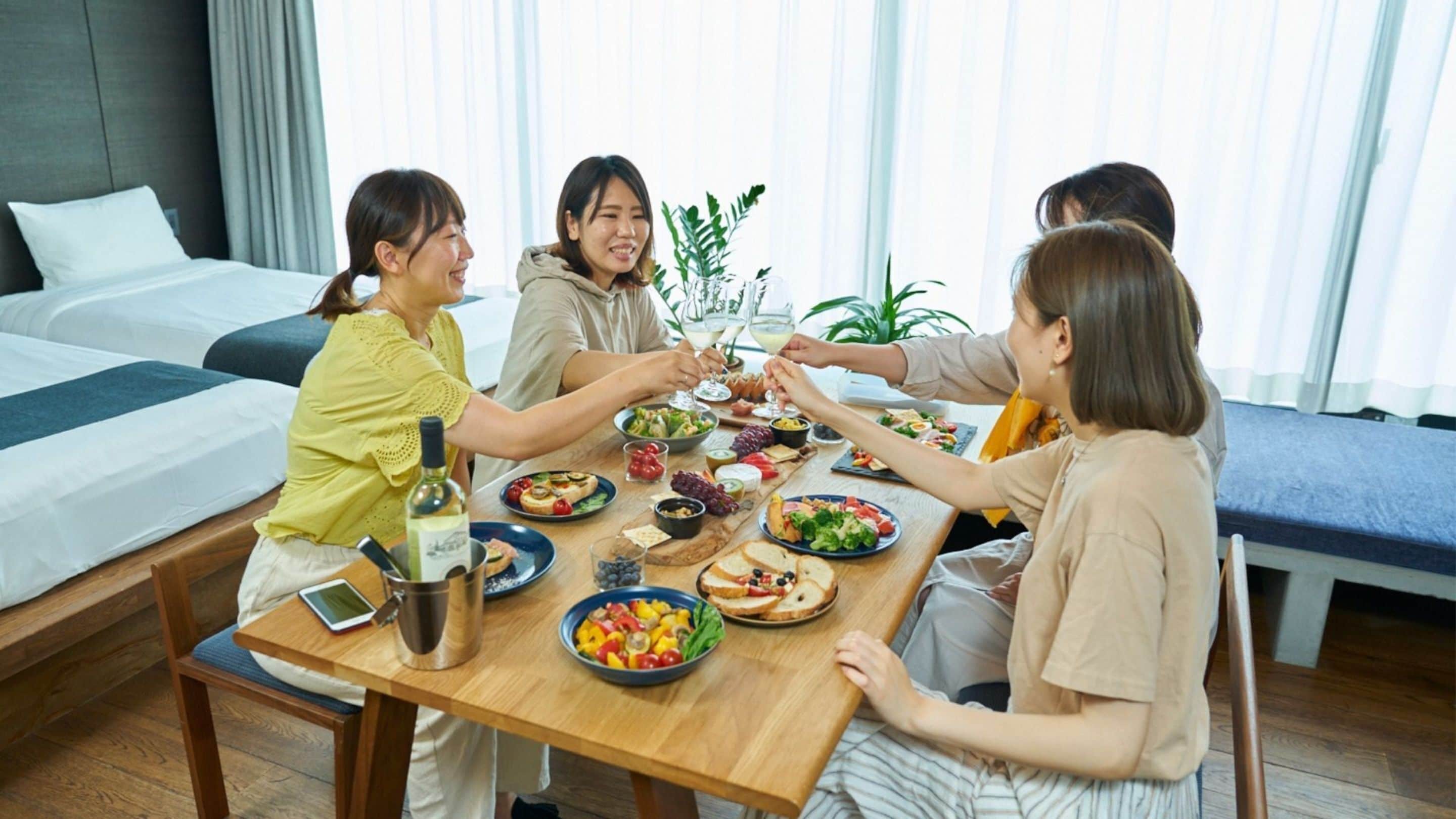 A group of people dining at a wooden table in a hotel room with a bed and curtains in the background.