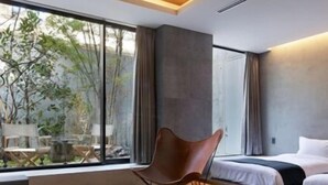 Ein modernes Hotelzimmer mit einem Glastisch, einem braunen Sessel, einem Bett und Blick auf Grünfläche.