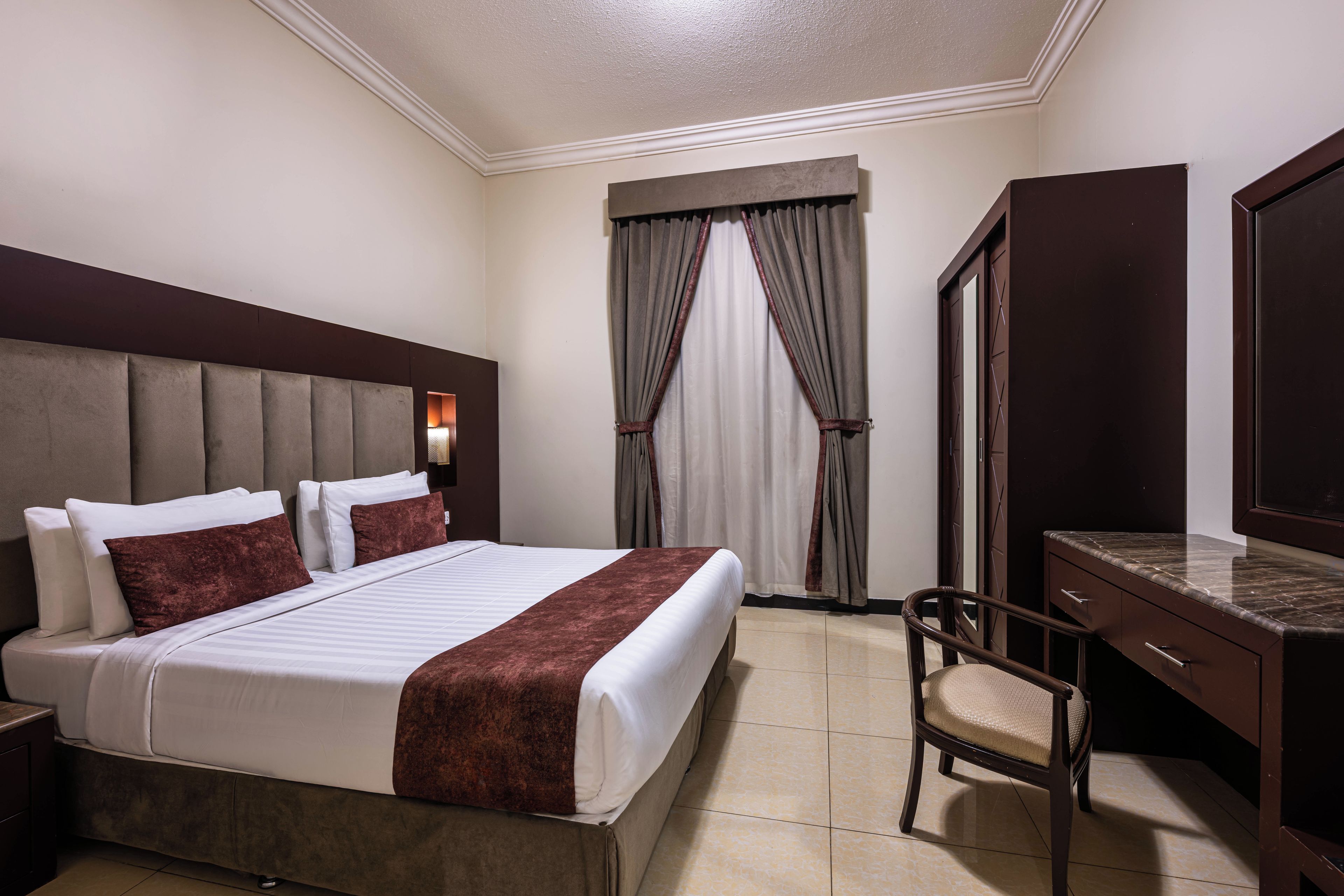 A hotel room with a large bed, a desk, a chair, a wardrobe, and a window with curtains.