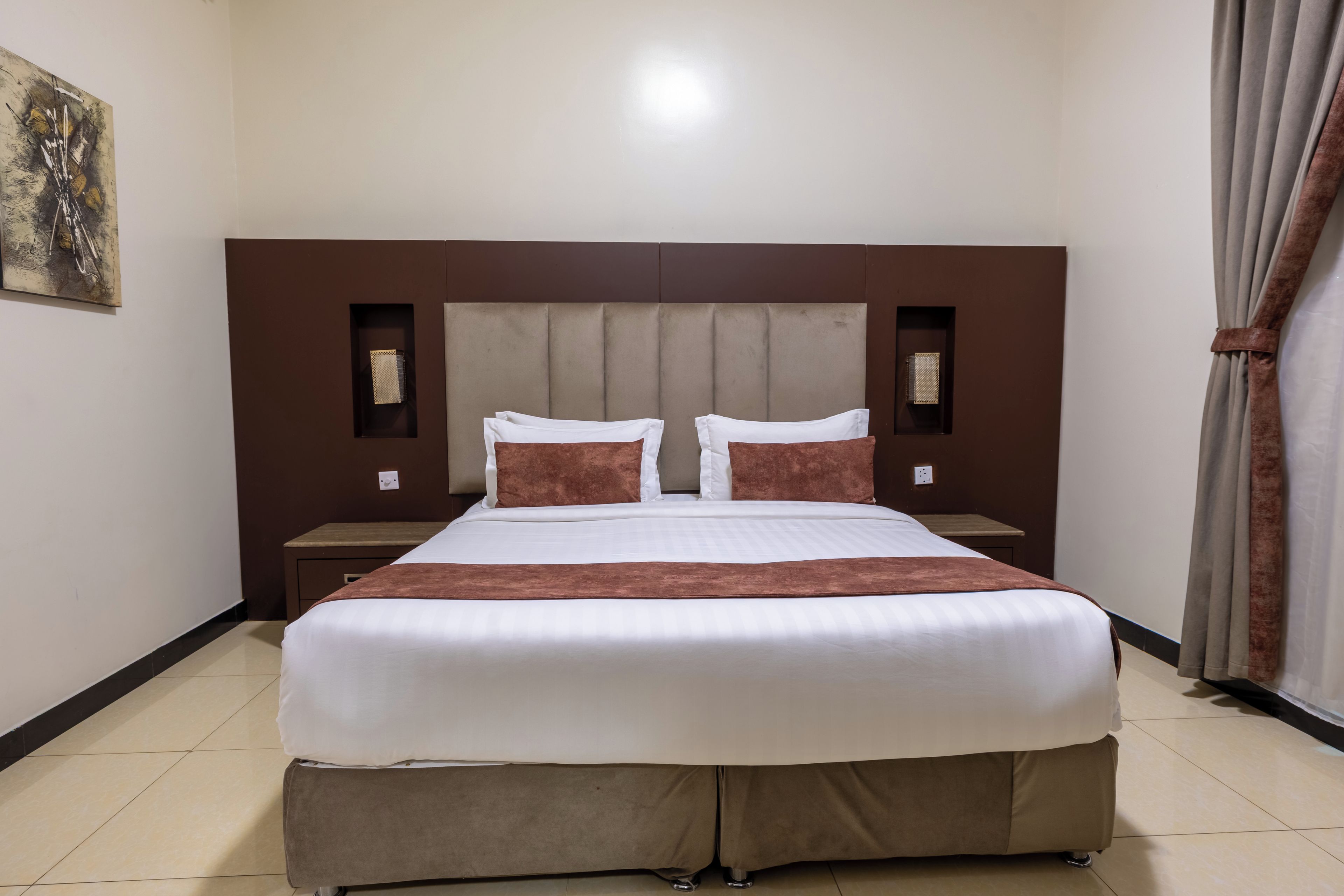 A hotel room with a large bed, two bedside tables, a headboard, and a painting on the wall.