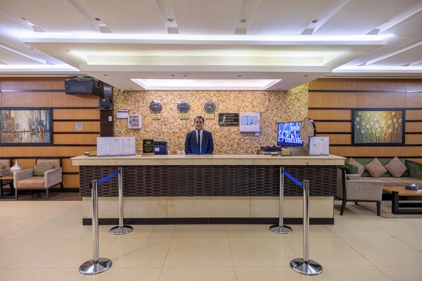 A hotel reception desk with a receptionist, a clock, a digital screen, and a QR code. - Al Muhaidb Residence Al ASKARY - Economic (Riyadh)
