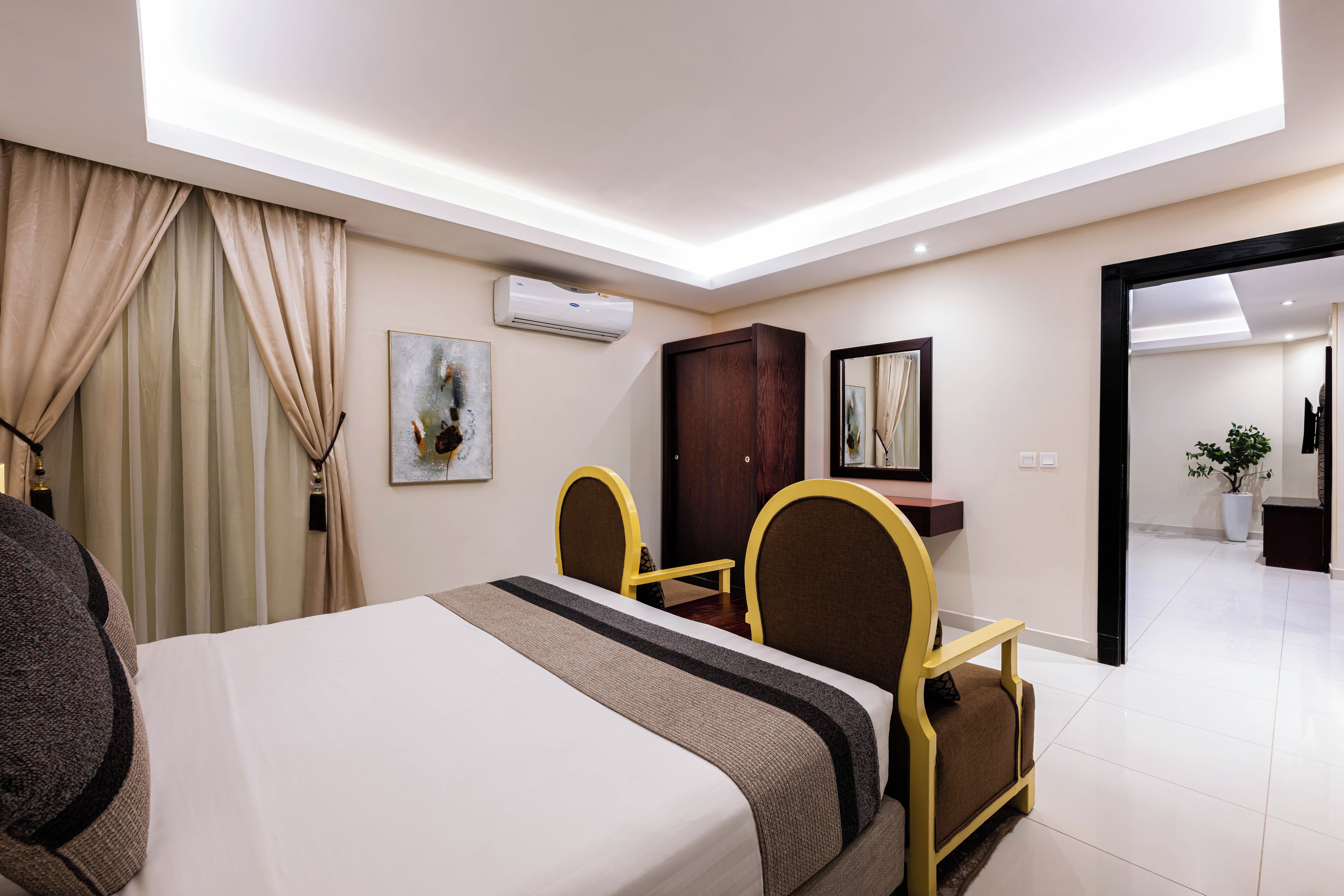 A hotel room with a bed, a desk, a chair, an air conditioning unit, a painting, and a mirror.