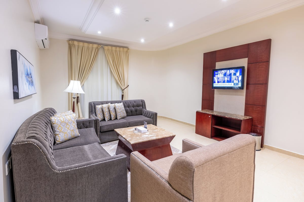 A modern living room with a grey sofa, beige armchair, and a coffee table. A flat-screen TV is mounted on the wall, and there is a lamp on a side table.