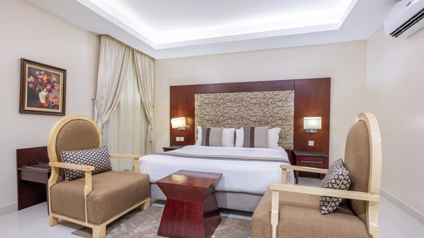 A hotel room with a large bed, two armchairs, a small wooden table, and a nightstand with a lamp. - Al Muhaidb Residence Al ASKARY - Economic (Riyadh)