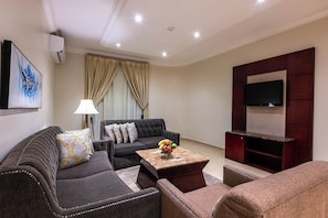 A modern living room with a sofa set, a coffee table with fruit, a TV mounted on a wooden panel, and a painting on the wall.