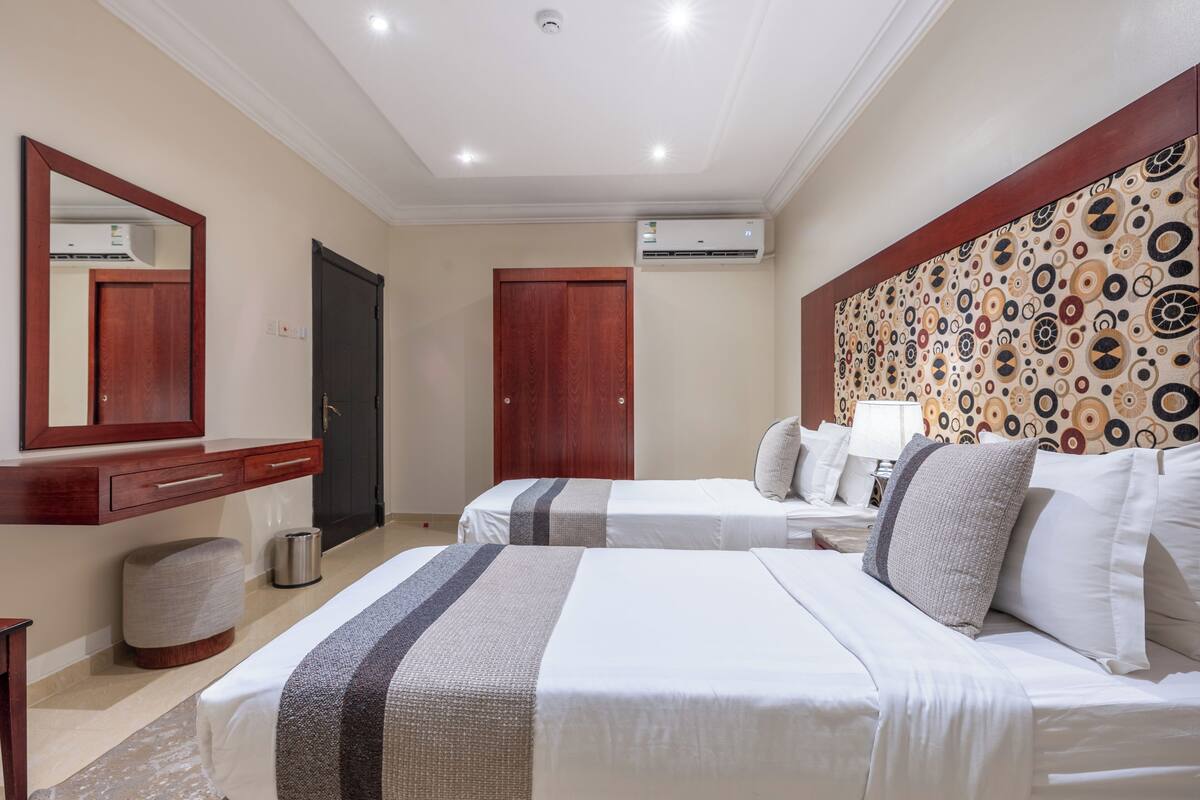A hotel room with two beds, a wooden wardrobe, a mirror, an air conditioning unit, and a patterned headboard.