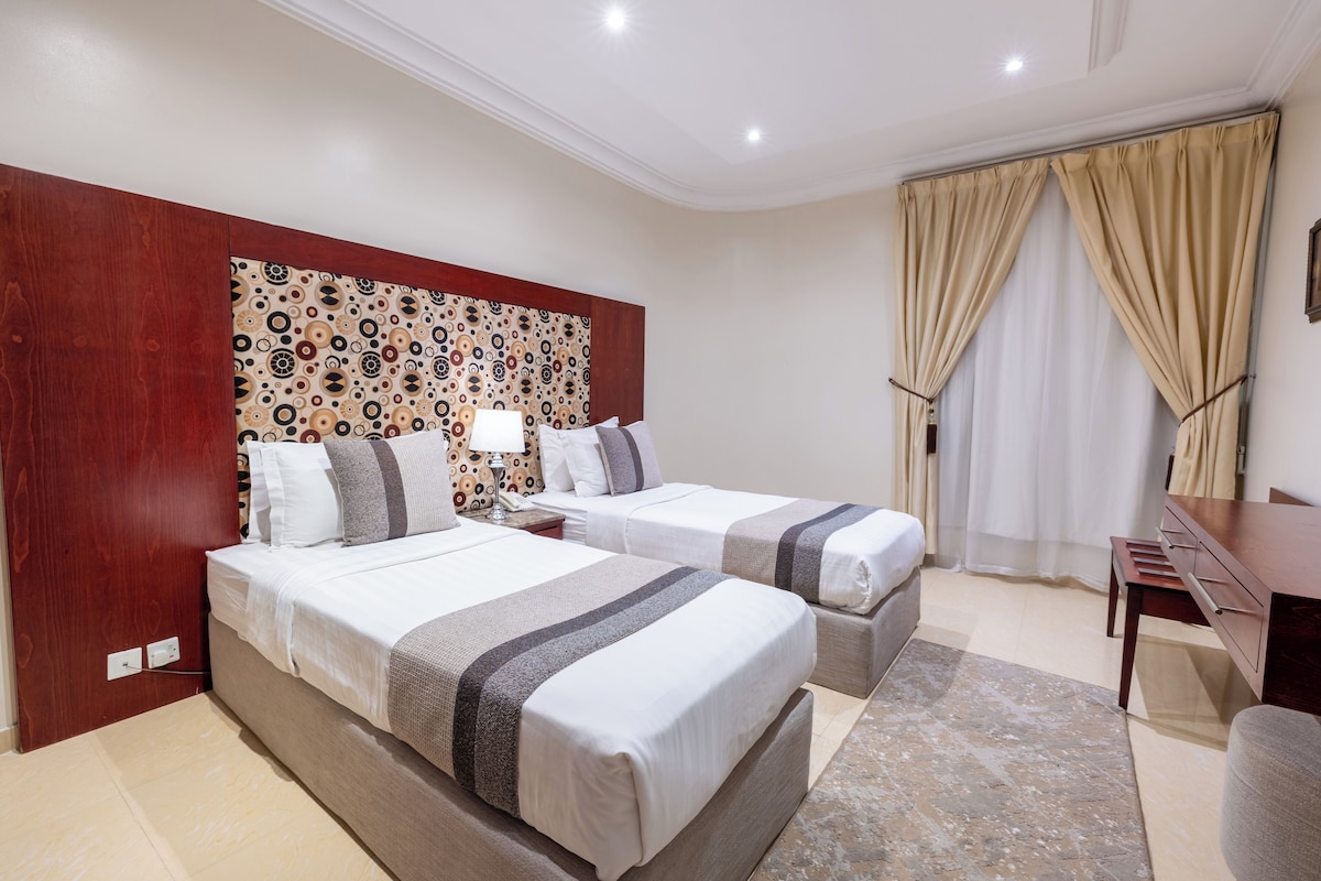 Habitación de hotel con dos camas, cabecera de madera, escritorio y un gran ventanal con cortinas.