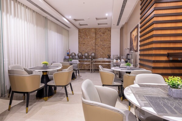 A modern dining area with round tables and chairs, a kitchen in the background, and a wooden accent wall. - Al Muhaidb Residence Al Malaz 5 - Economic (Riyadh)
