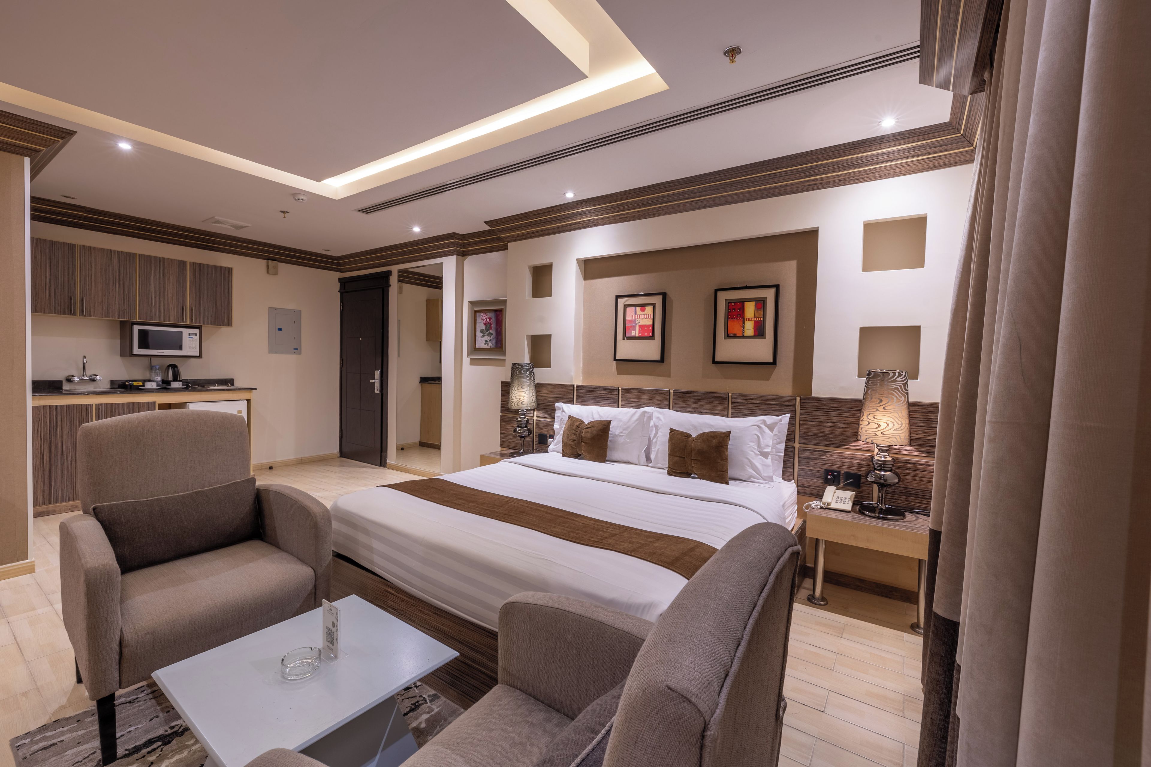 A hotel room with a bed, two armchairs, a small table, a desk with a lamp, a telephone, and artwork on the wall.