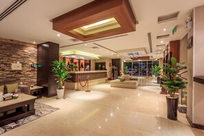 Un moderno hall de hotel con una recepción, un área de descanso y una escalera que conduce a un nivel superior.