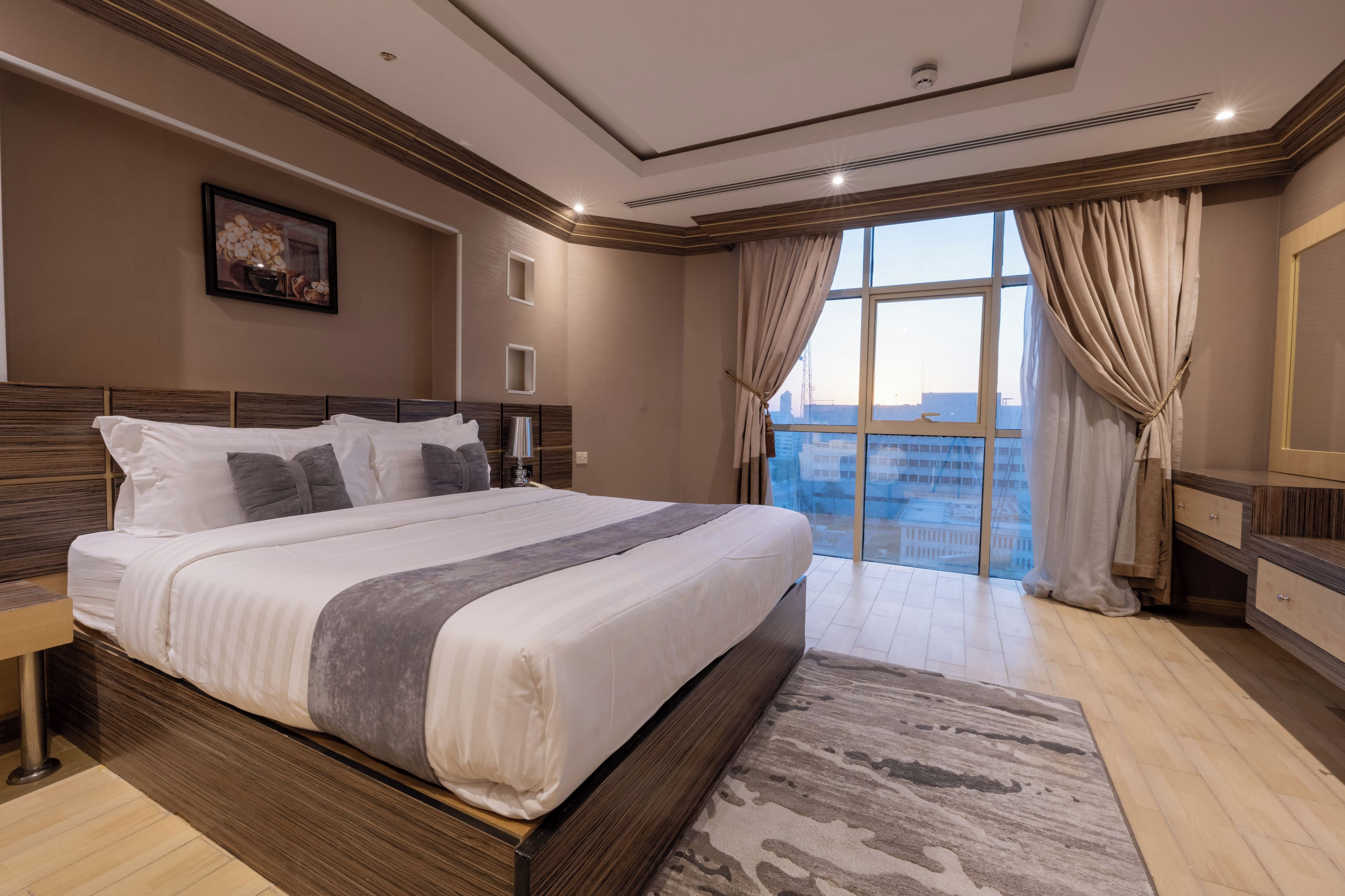 A modern bedroom with a large bed, a nightstand, a bedside table, a window with curtains, and a view of the cityscape.