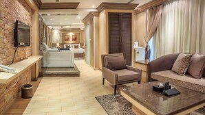 Una moderna camera d'albergo con area soggiorno, divano, tavolino e televisore montato su una parete in mattoni. Si intravedono la cucina e il bagno.
