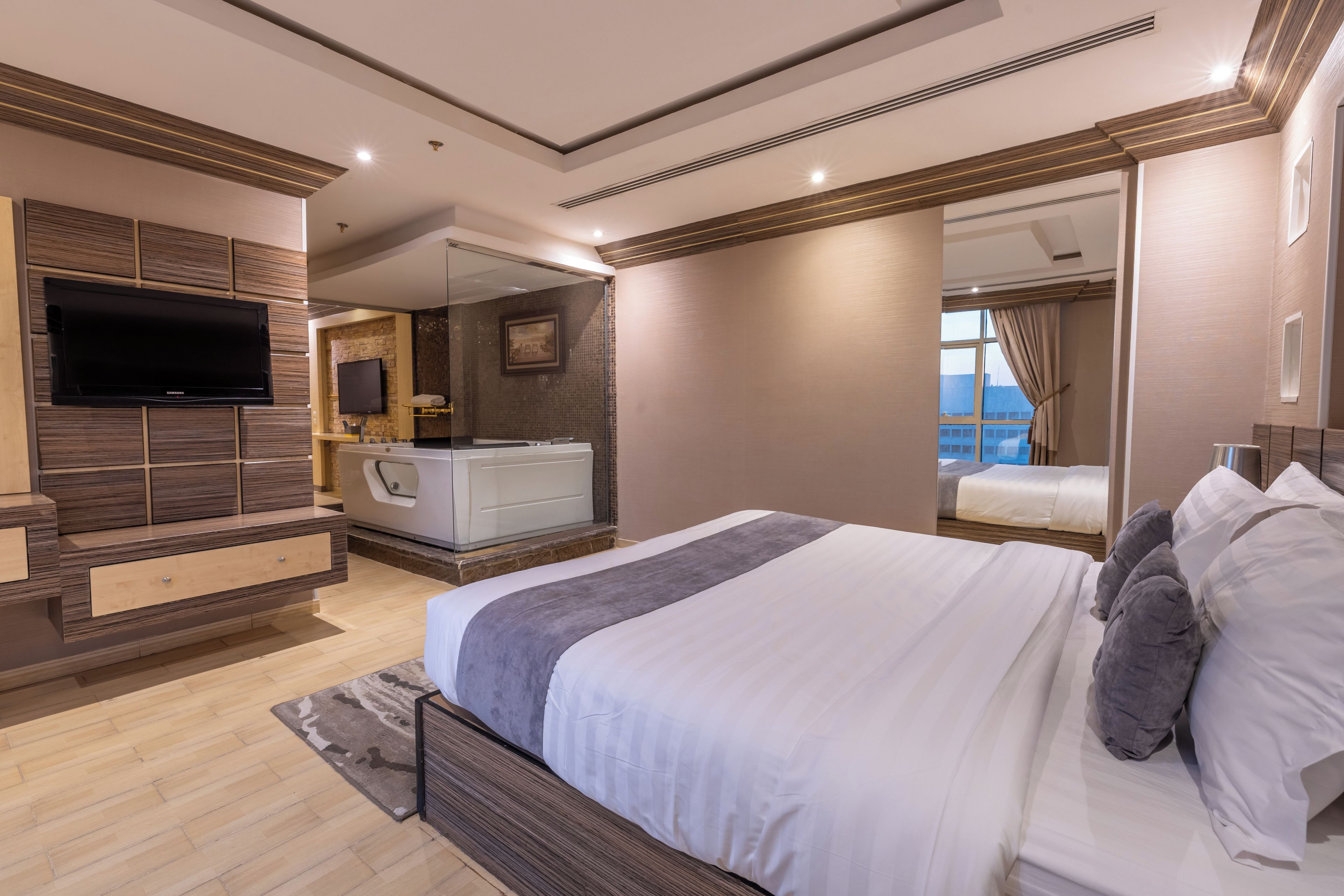 A modern bedroom with a large bed, a flat-screen TV, a vanity with a mirror, and a view of the city through a window.
