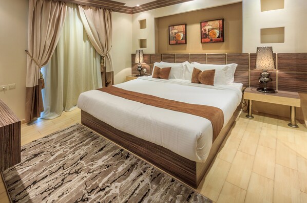 Habitación de hotel con una cama grande, cabecera de madera, mesita de noche con lámpara y un ventanal con cortinas.