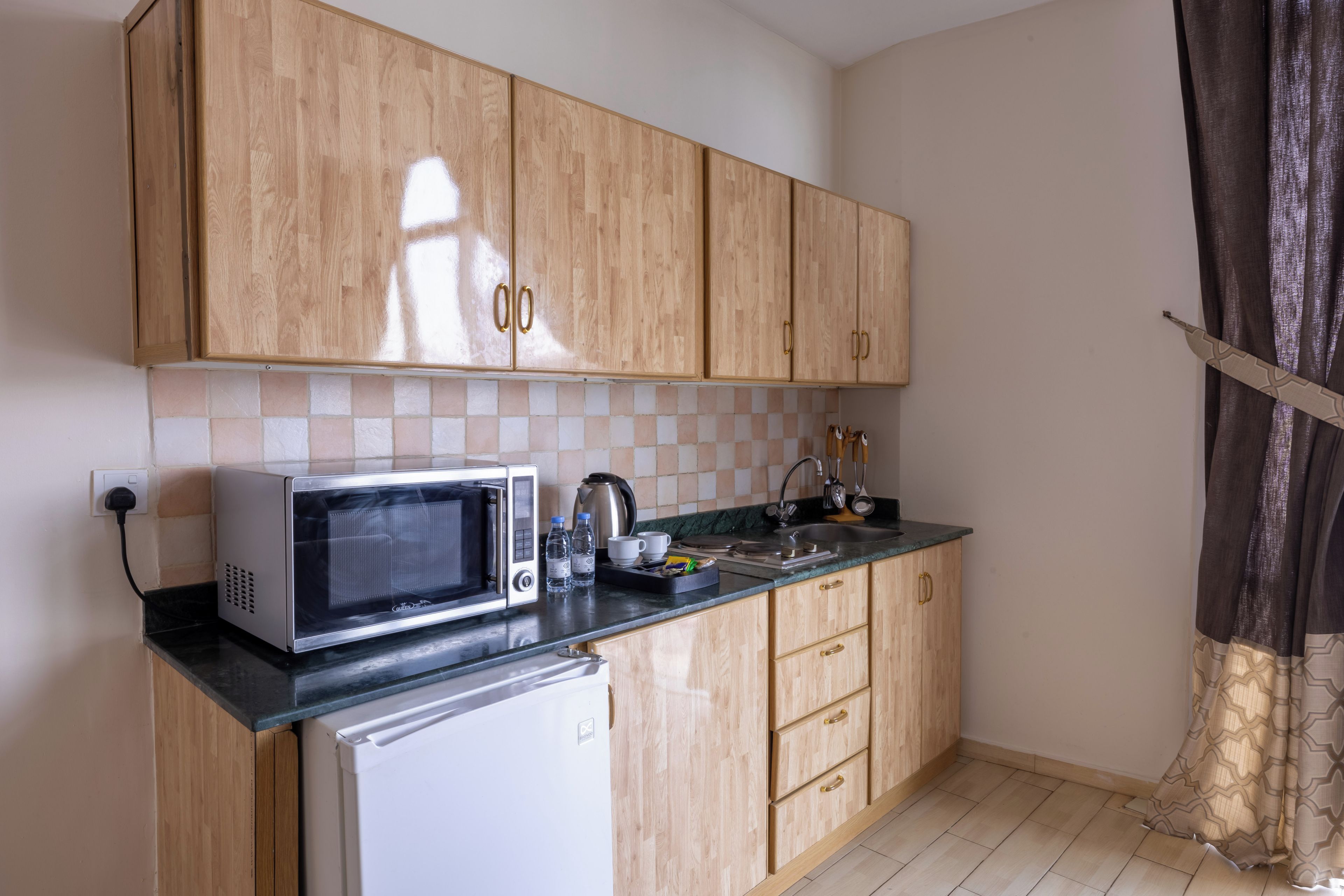 A compact kitchen with wooden cabinets, a microwave, a kettle, and a refrigerator.