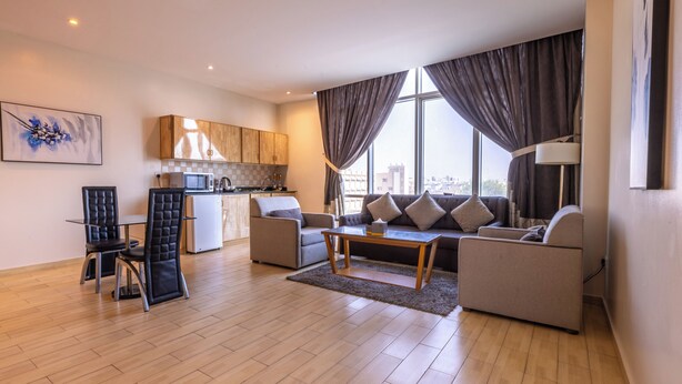A modern living room with a sofa, chairs, a coffee table, a kitchen area, and large windows with curtains.