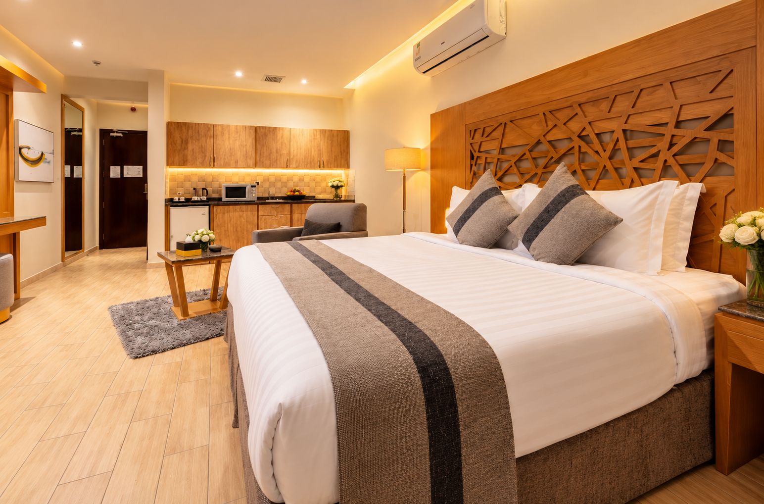 A modern hotel room with a large bed, a wooden headboard, a small kitchenette, and a separate seating area.