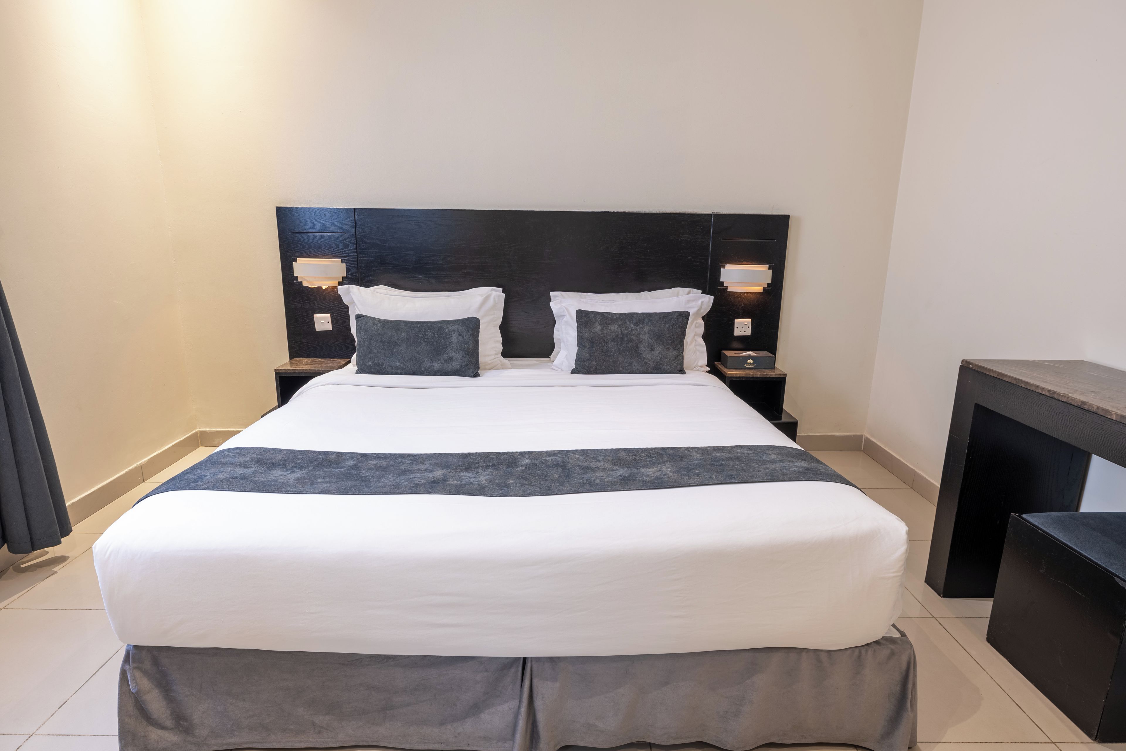 A neatly made bed with a white and gray bedspread, two pillows, and a matching throw. There are two bedside tables with lamps, a small bedside table with a box, and a wooden bench.