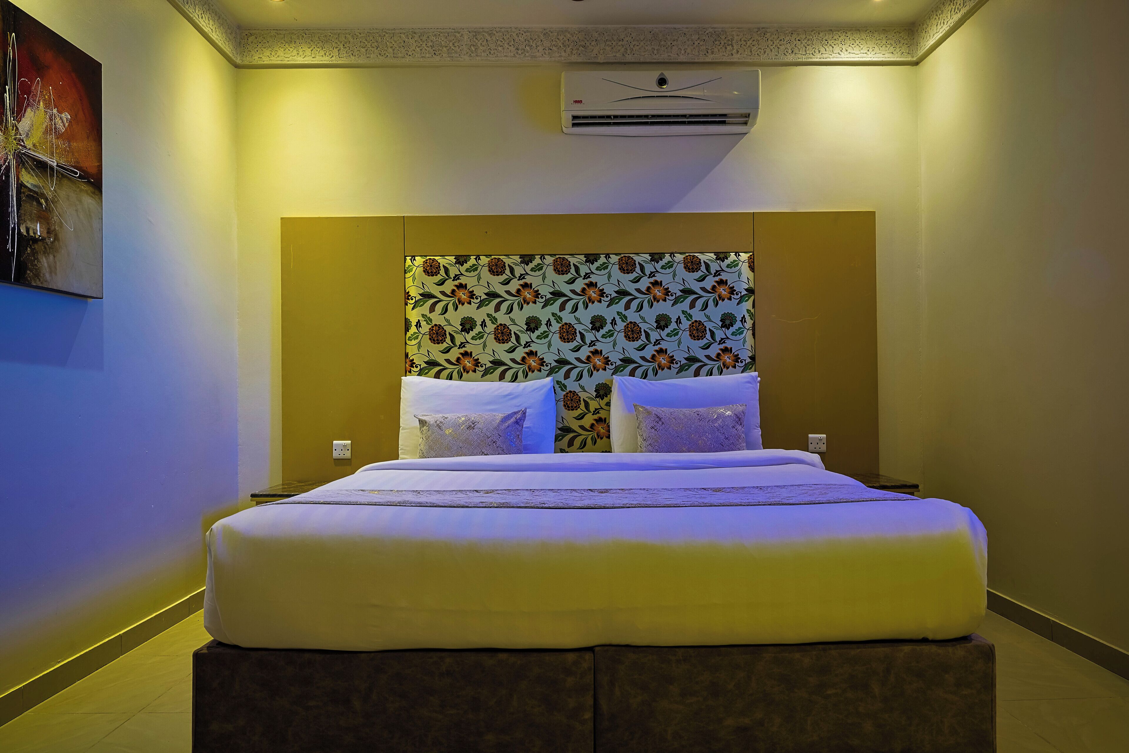 A hotel room with a large bed, a headboard with floral wallpaper, and an air conditioning unit on the wall.