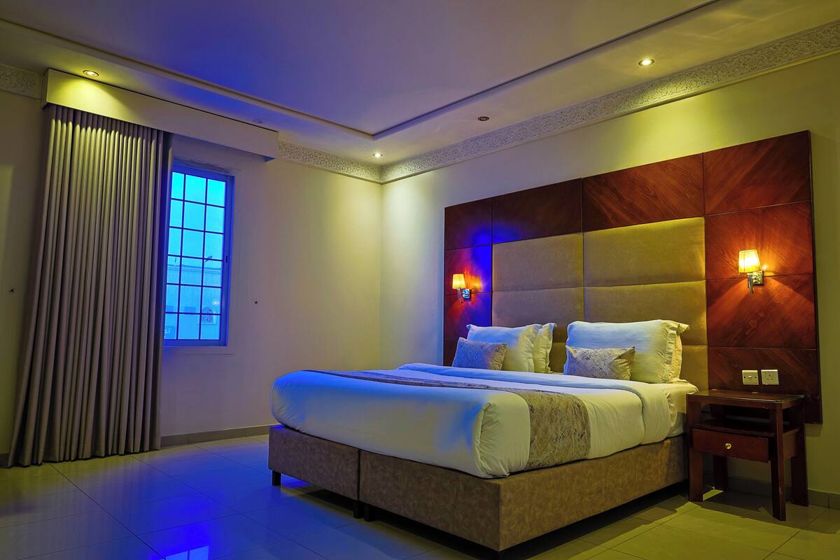 A modern hotel room with a large bed, a nightstand, a window with curtains, and a wall-mounted light fixture.