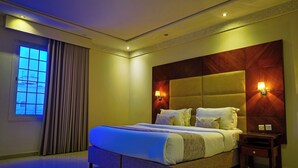 A hotel room with a large bed, a nightstand, a window with curtains, and a wall-mounted light fixture. - DAR NEYAARA SERVICED APARTMENTS - Economic (Riyadh)