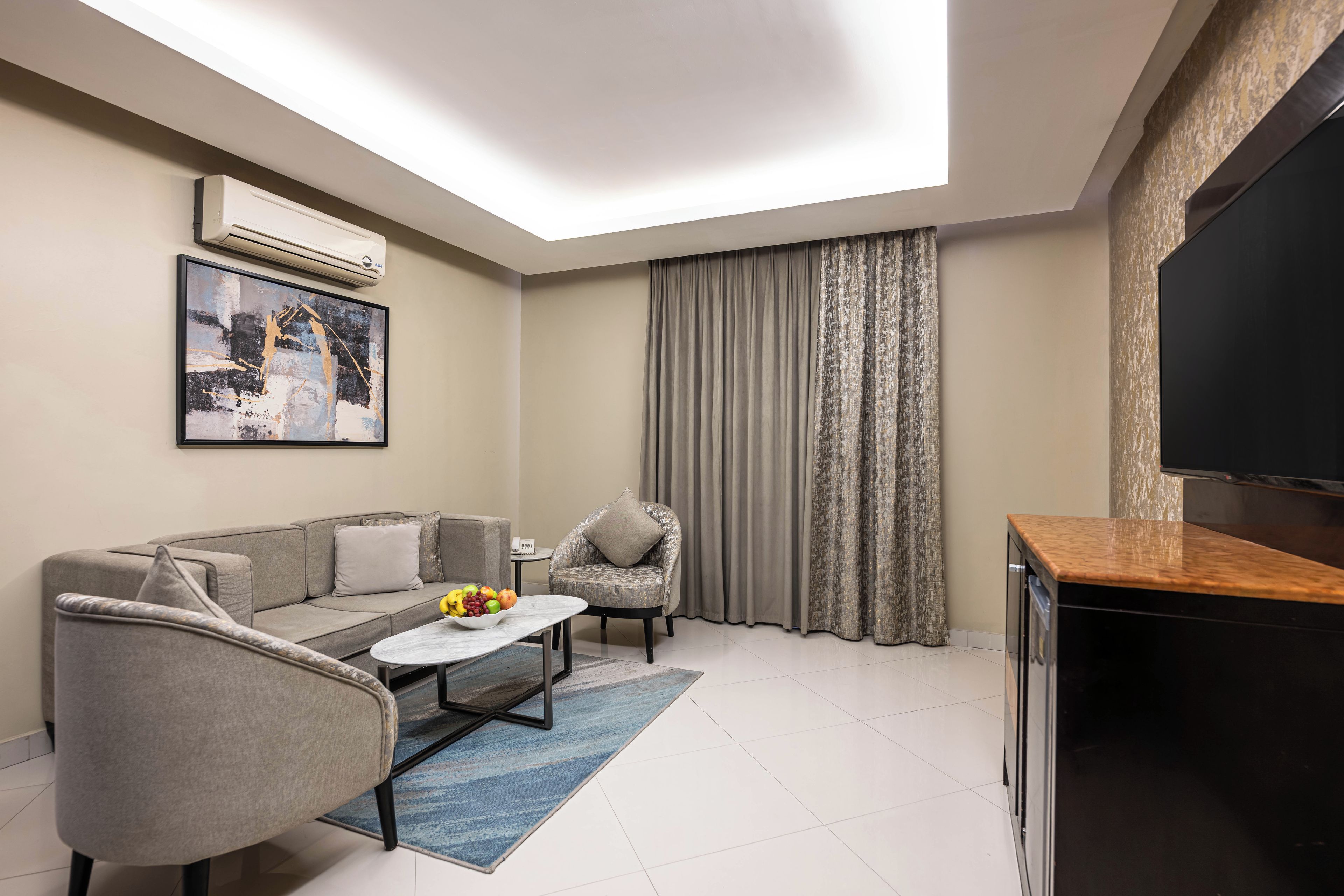 Una sala de estar moderna con un sofá gris, una mesita redonda con fruta, un sillón gris, un televisor y un amplio ventanal con cortinas.