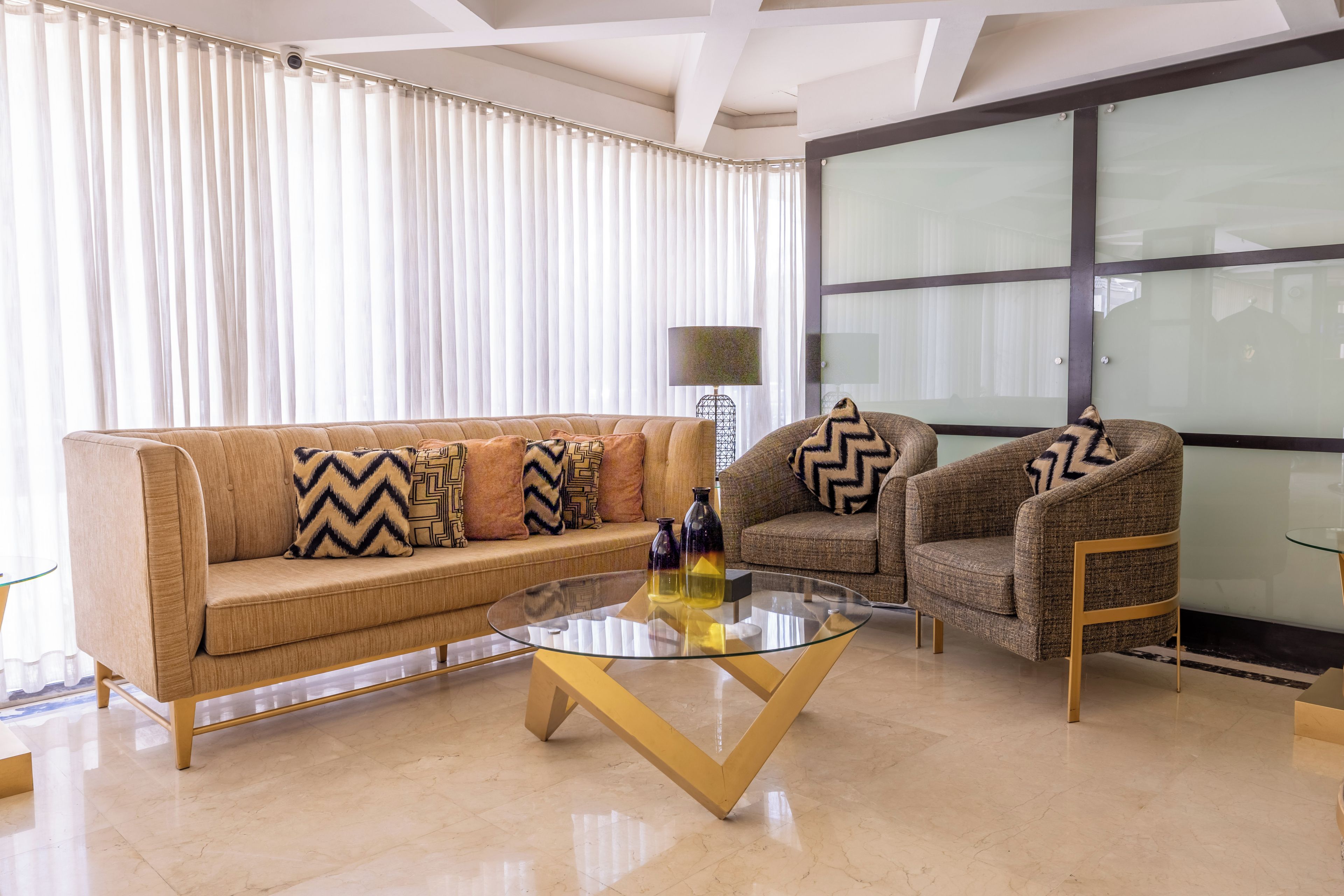 A modern living room with a beige sofa, two armchairs, a glass coffee table, and a large window with curtains.