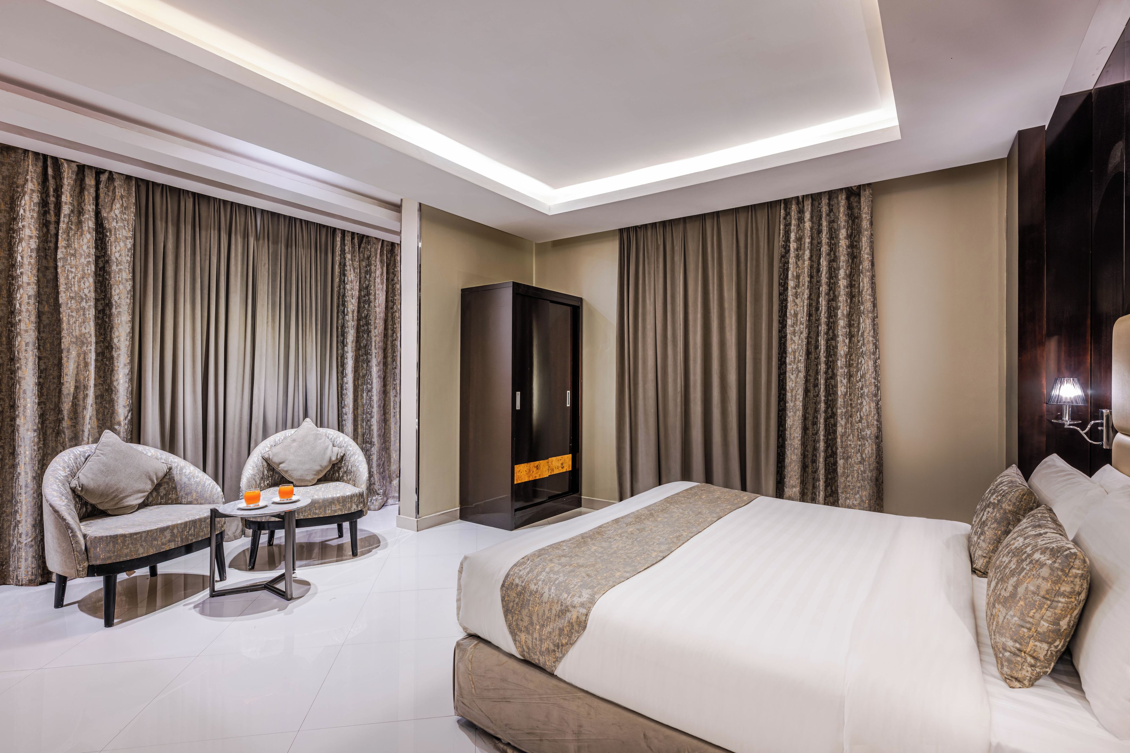 Ein Hotelzimmer mit einem Bett, zwei Sesseln, einem kleinen Tisch mit Getränken, einem Kleiderschrank und Vorhängen.
