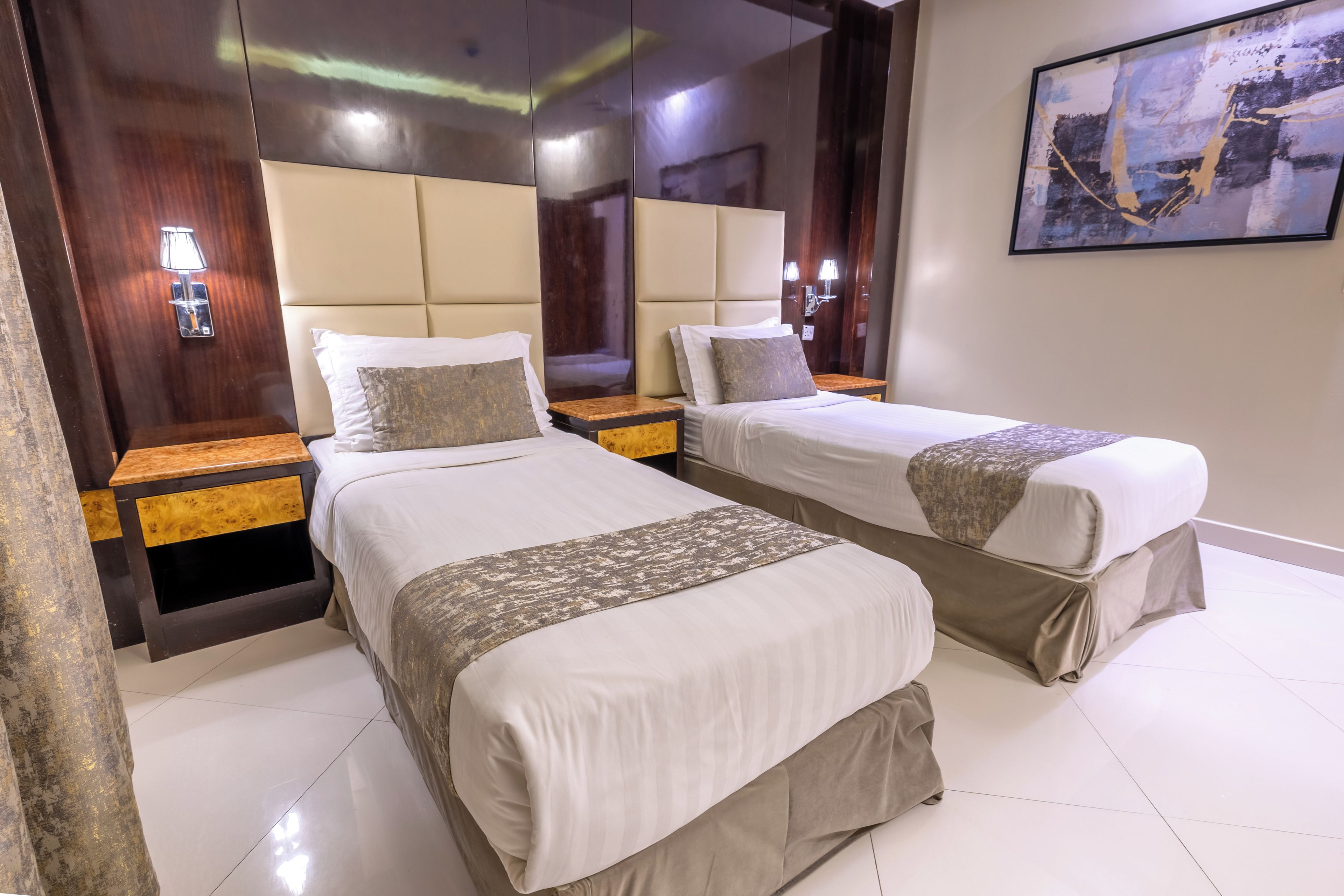 A hotel room with two beds, wooden headboards, a nightstand with a lamp, and a painting on the wall.