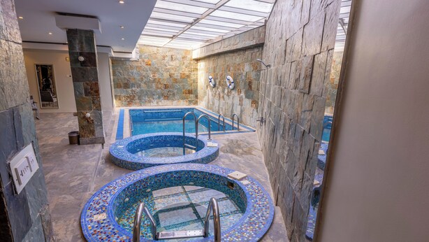 A spa area with two circular hot tubs and a rectangular pool, surrounded by stone walls and a glass ceiling.
