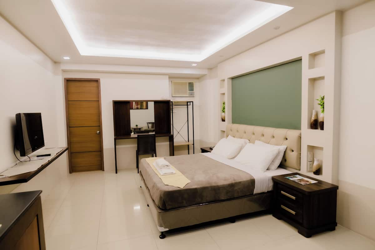 Quarto de hotel moderno com cama, mesas de cabeceira, escrivaninha, televisão e luminária no teto.