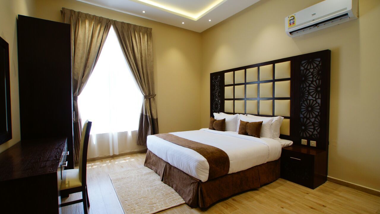 A hotel room with a bed, a desk, a chair, a window with curtains, an air conditioning unit, and a decorative headboard.