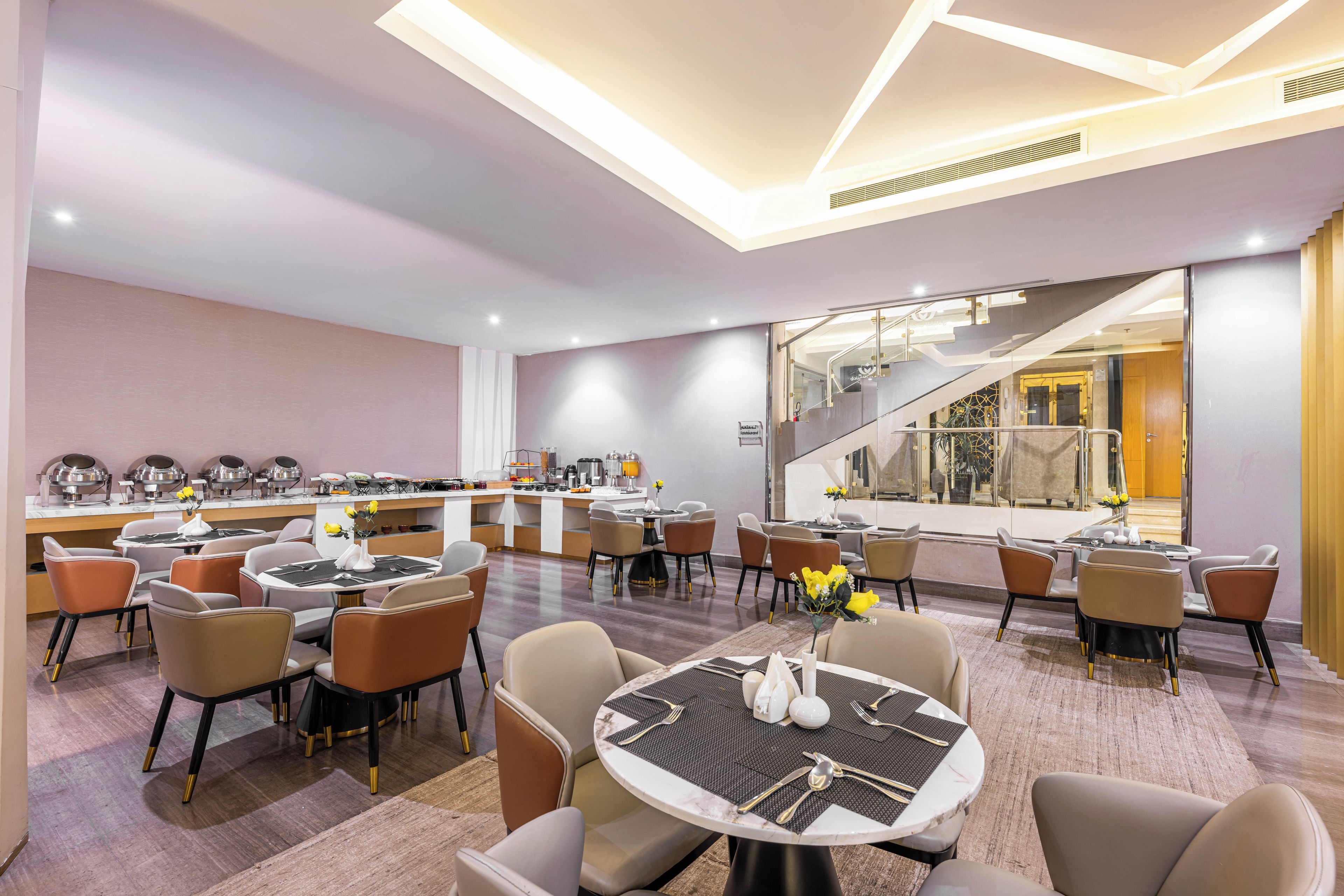 Un área de comedor de hotel moderna con varias mesas y sillas, un bar y una escalera que lleva a un nivel superior.