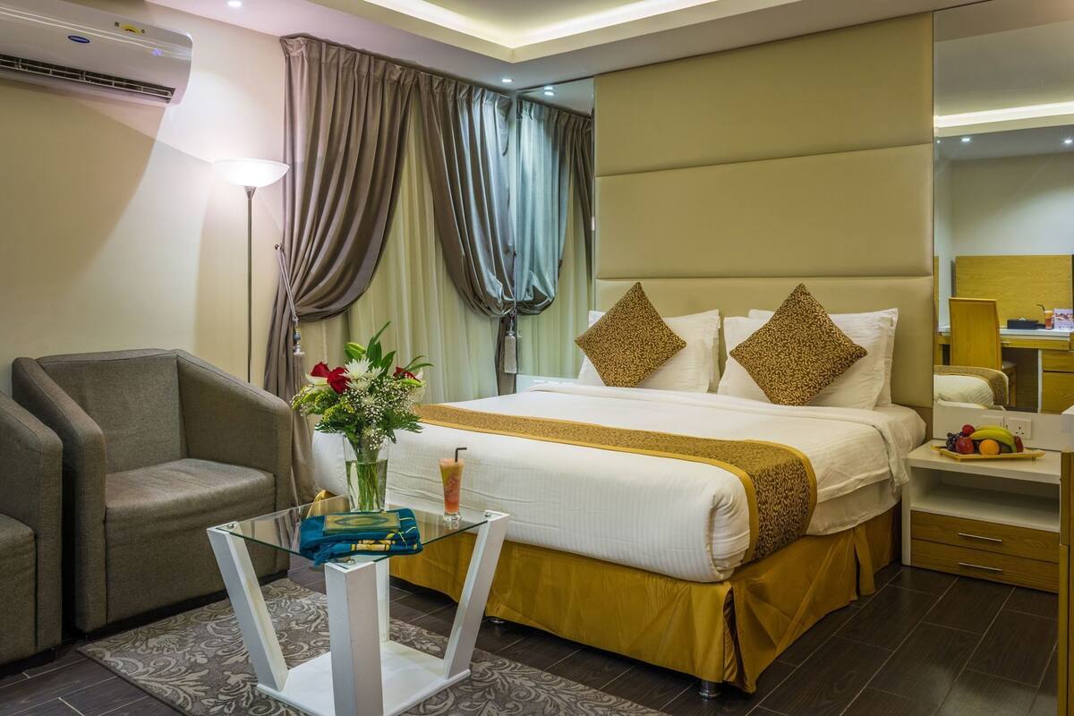 A hotel room with a bed, a chair, a desk, a lamp, a vase with flowers, and a fruit basket.