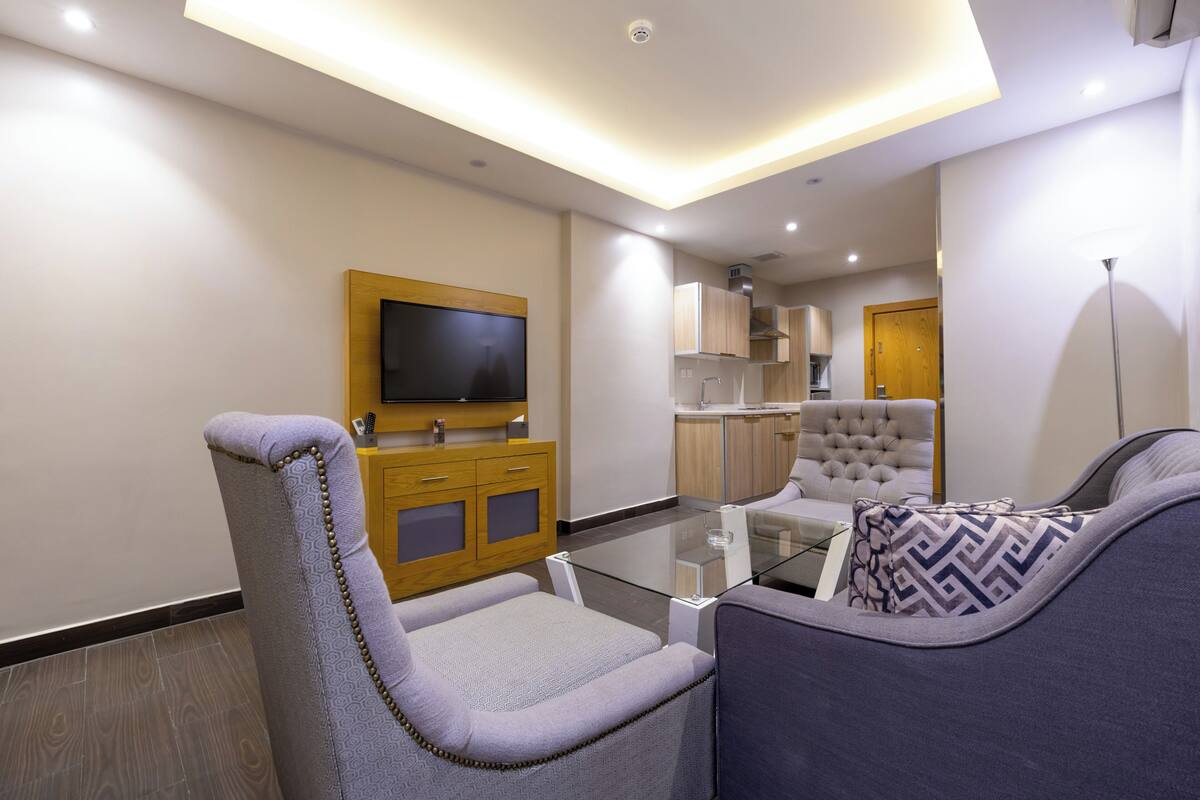 A modern living room with a glass coffee table, comfortable seating, a flat-screen TV mounted on a wooden cabinet, and a kitchen area in the background.